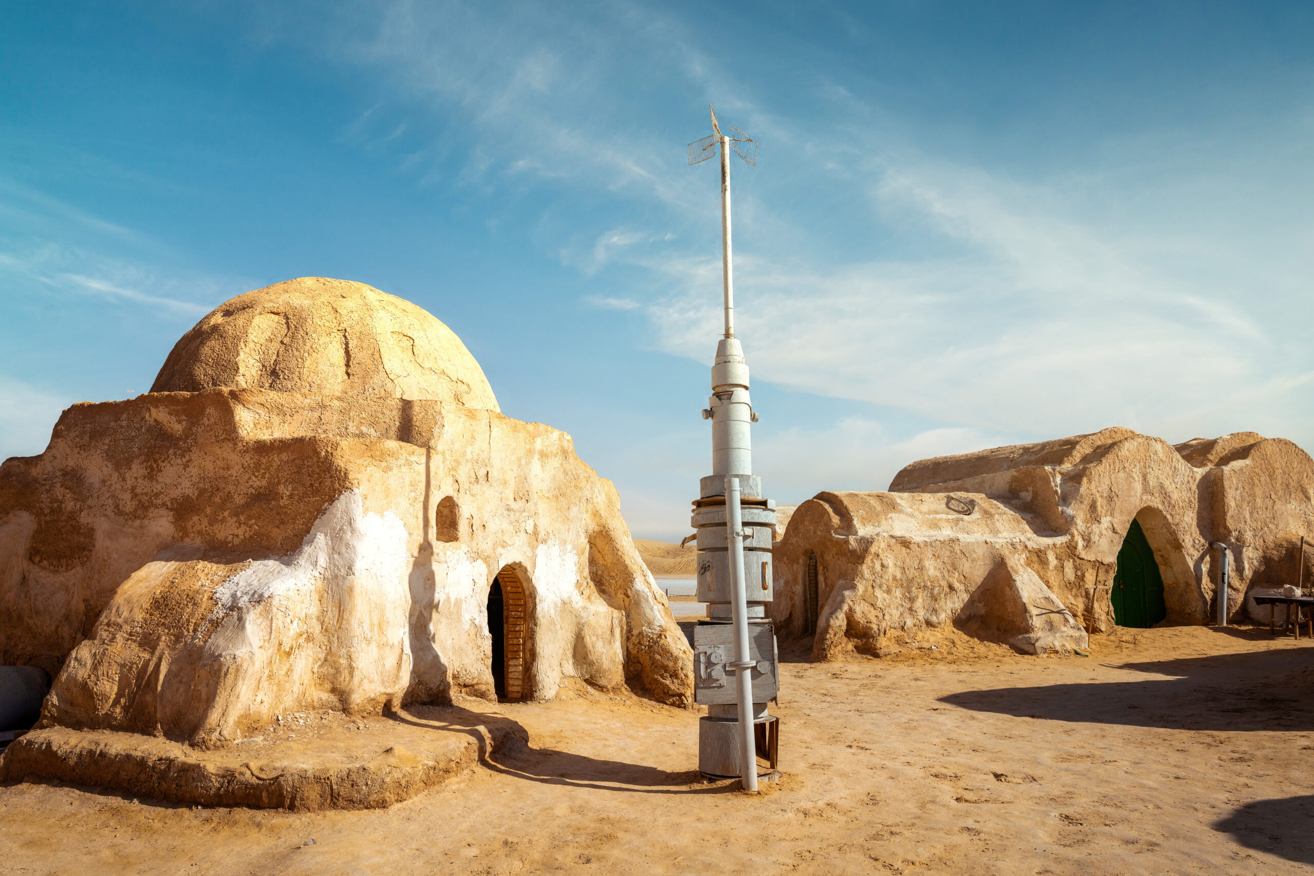 Desert village with sandy domed houses and a tall metal antenna against a blue sky.