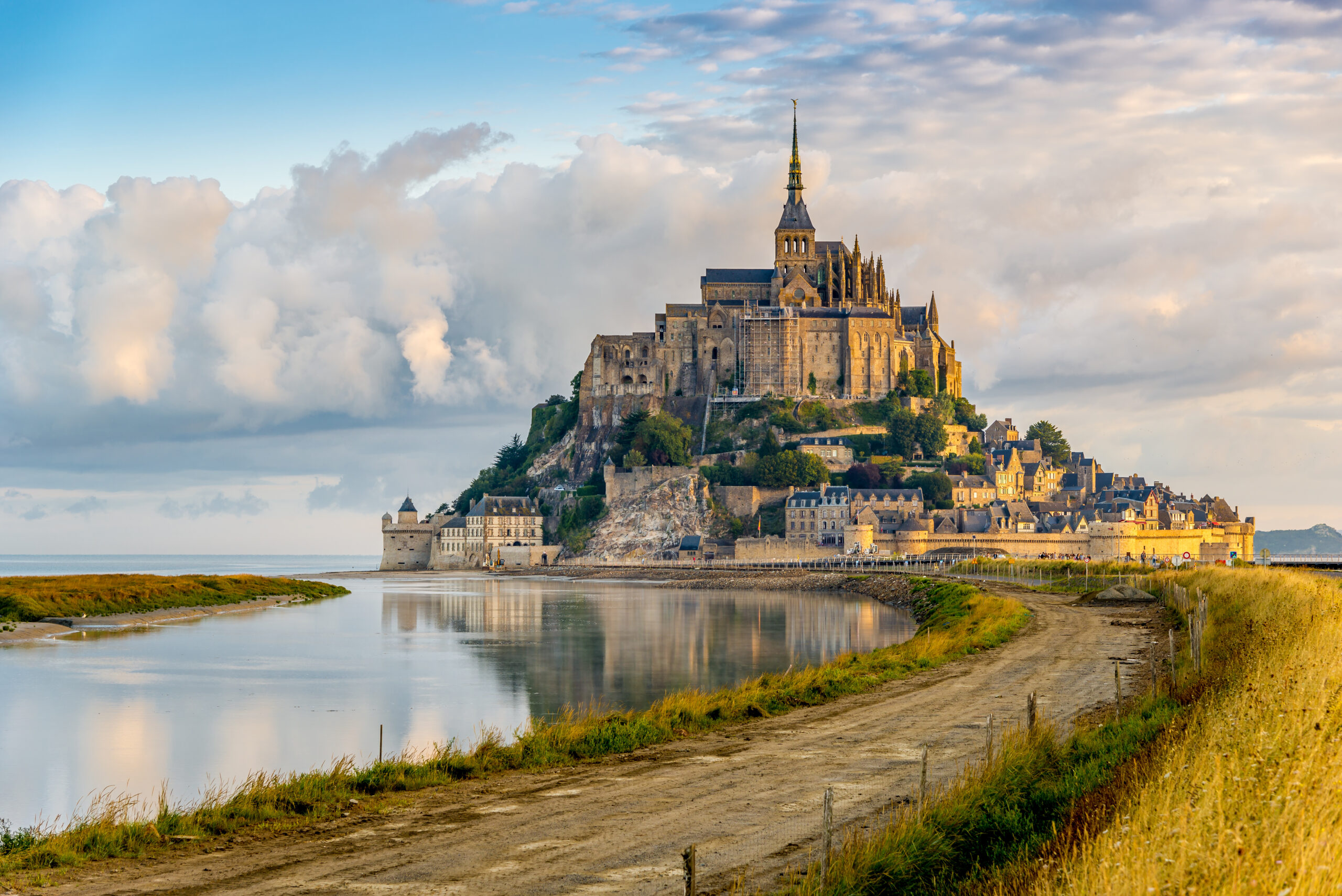 Mont Saint-Michel abbey perched on a rocky island with surrounding walls and village at low tide, under a blue sky with clouds nearby reflection in water