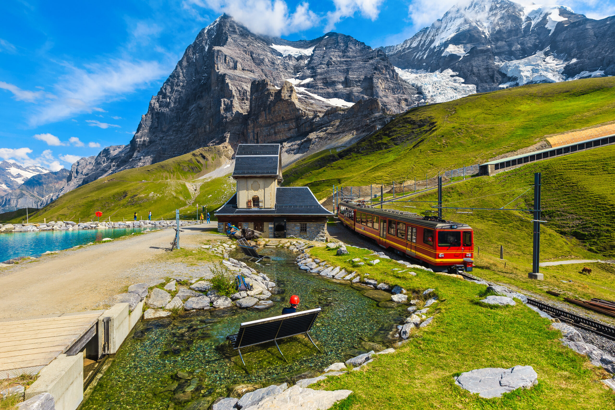Alpine landscape with snow‑capped peaks, green slopes, a red train beside a small stone station, and a clear stream in the foreground under a blue sky.