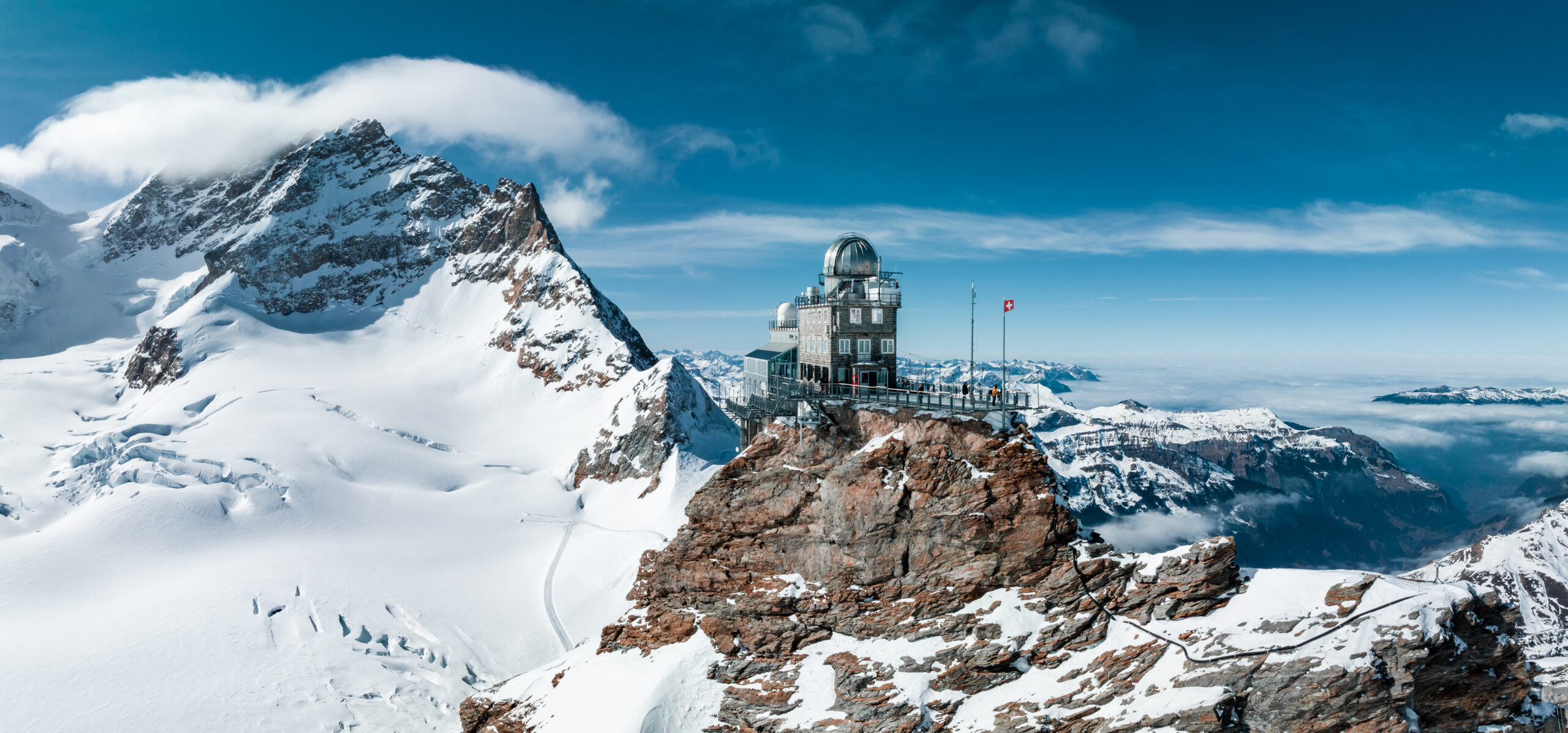 Mountain weather/observatory building on a rocky summit above snow, with a silver dome and a Swiss flag nearby.