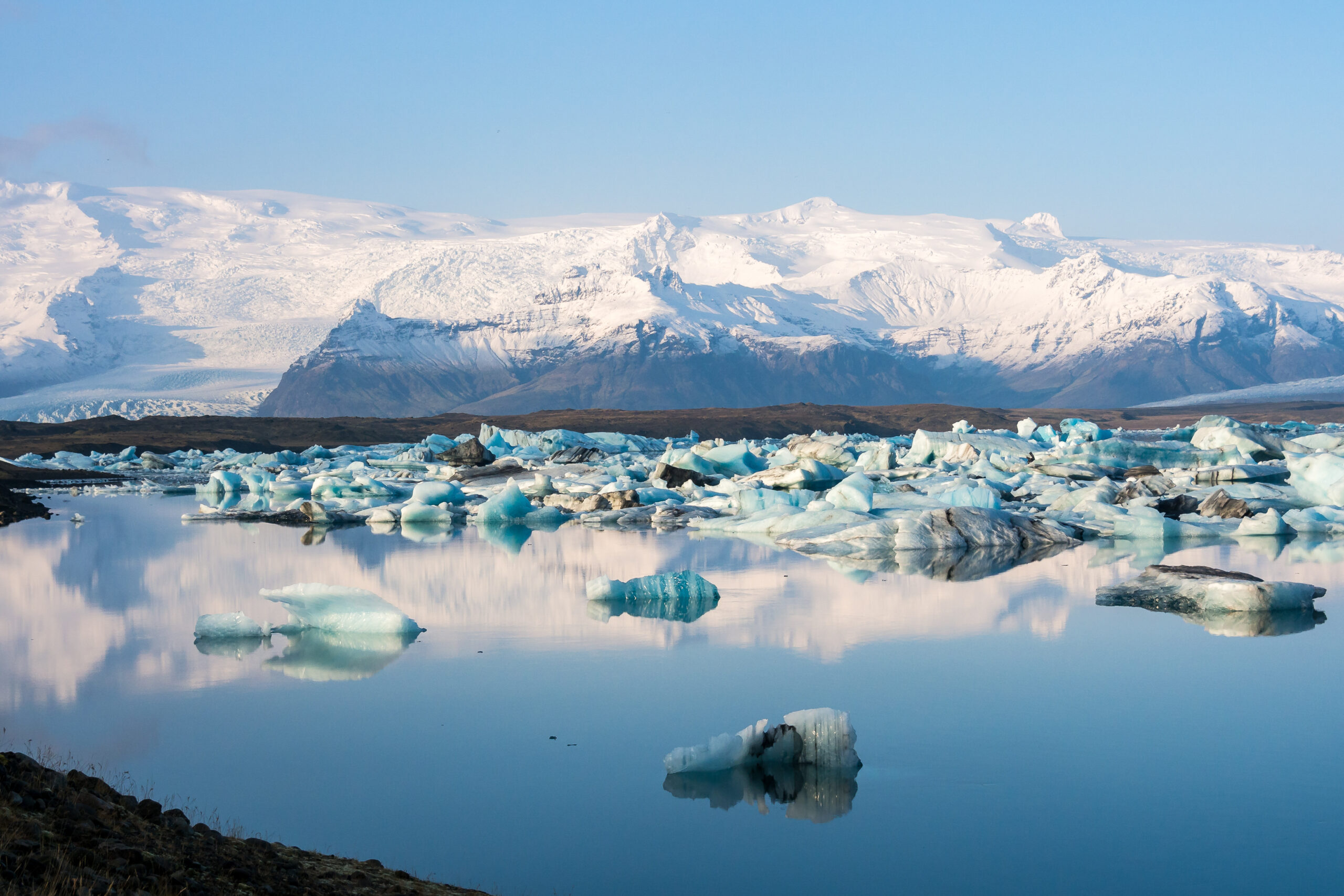 Snowy mountain glacier with blue ice chunks floating on a calm lagoon under a clear blue sky