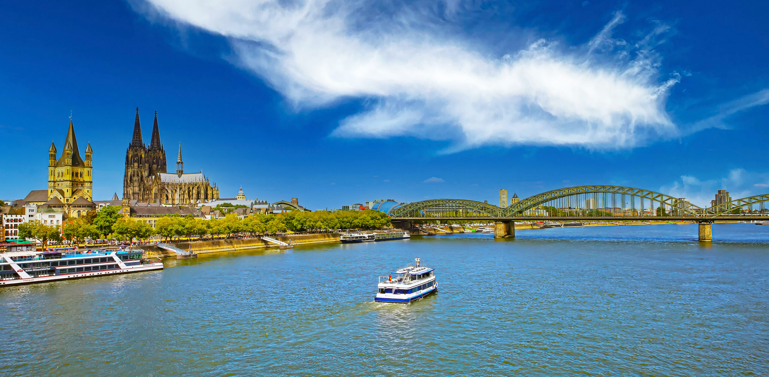 Cologne skyline along the Rhine with the Gothic cathedral and the Hohenzollern Bridge under a clear blue sky, boats on the water.