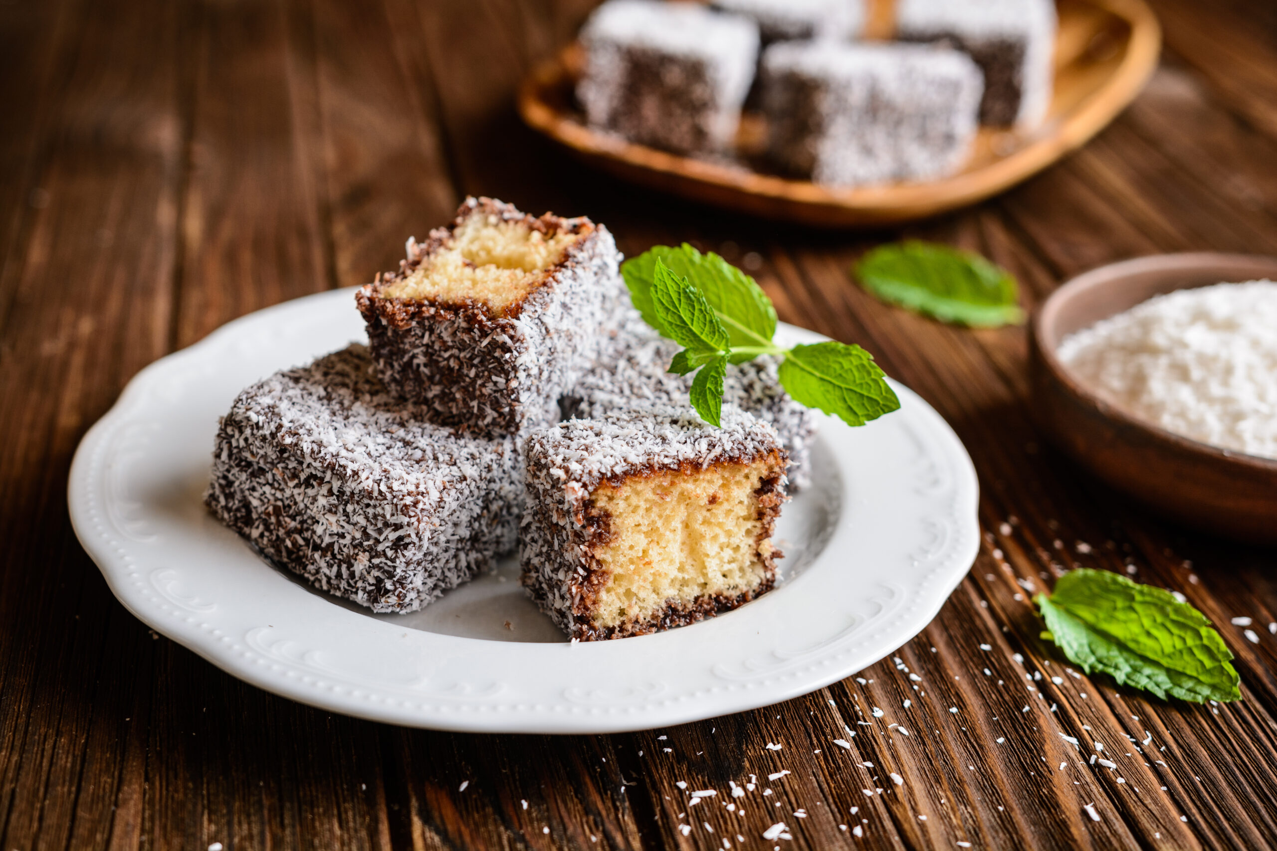 Coconut-coated vanilla cake squares on a white plate, garnished with fresh mint leaves.