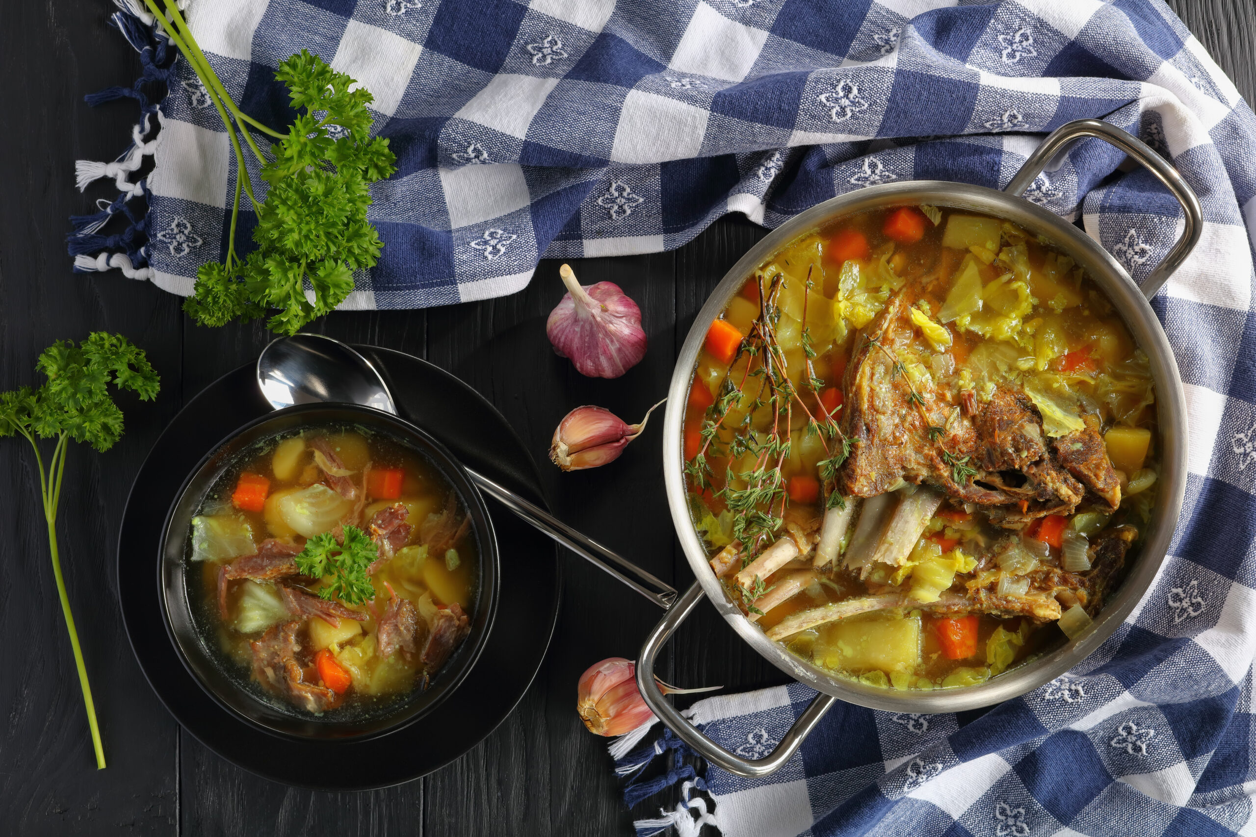 Pot of hearty vegetable soup with herbs, carrots and potatoes on a blue checkered cloth, garlic nearby.