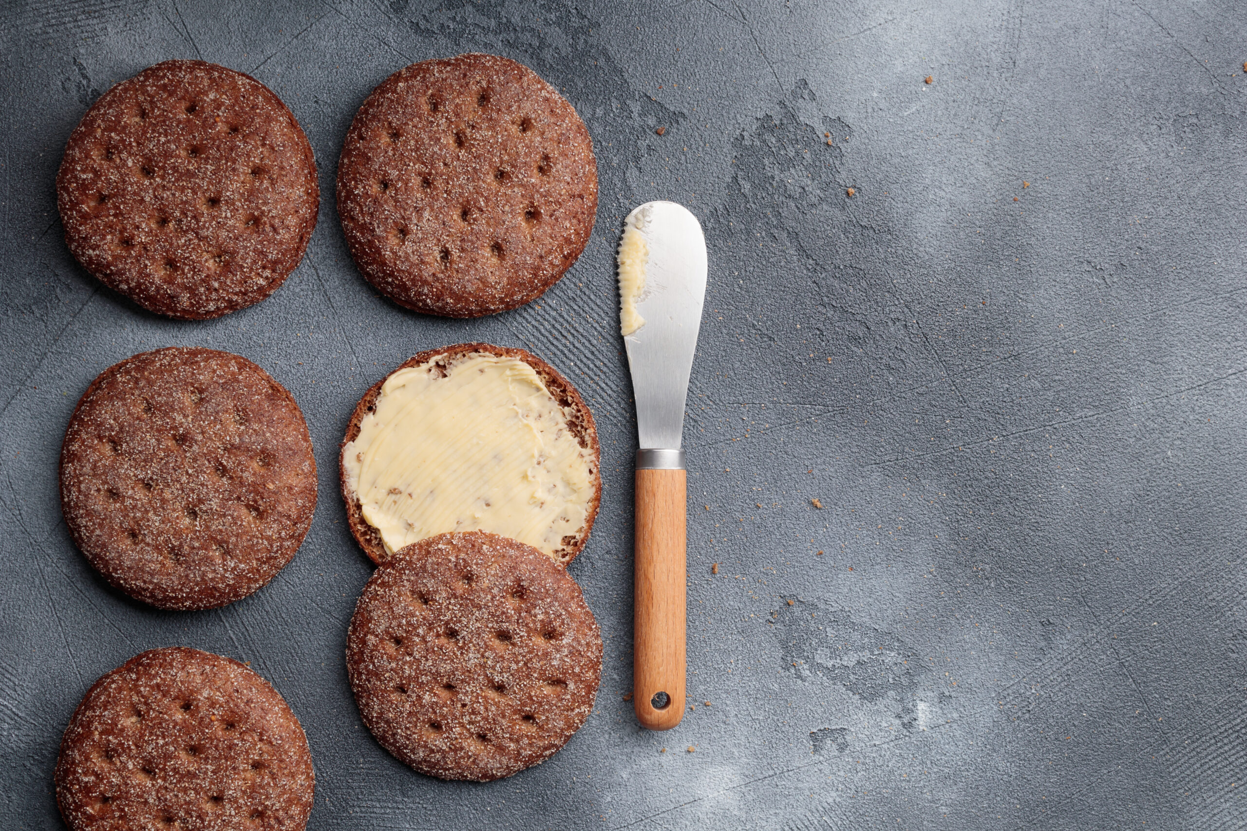 Several chocolate cookies dusted with sugar, one half-coated with butter spread using a butter knife on a gray surface