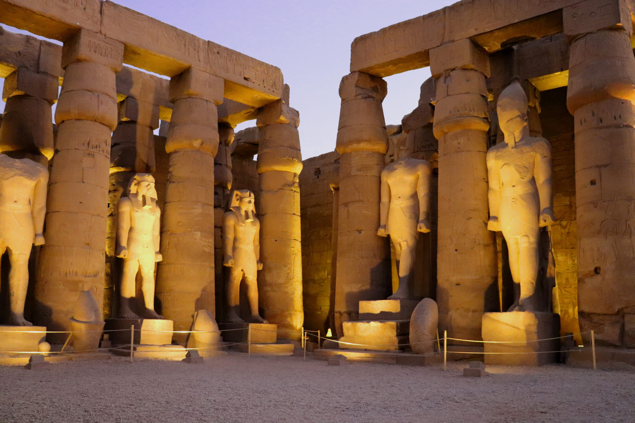 Massive ancient Egyptian temple facade with sandstone columns and colossal statues, illuminated at dusk.