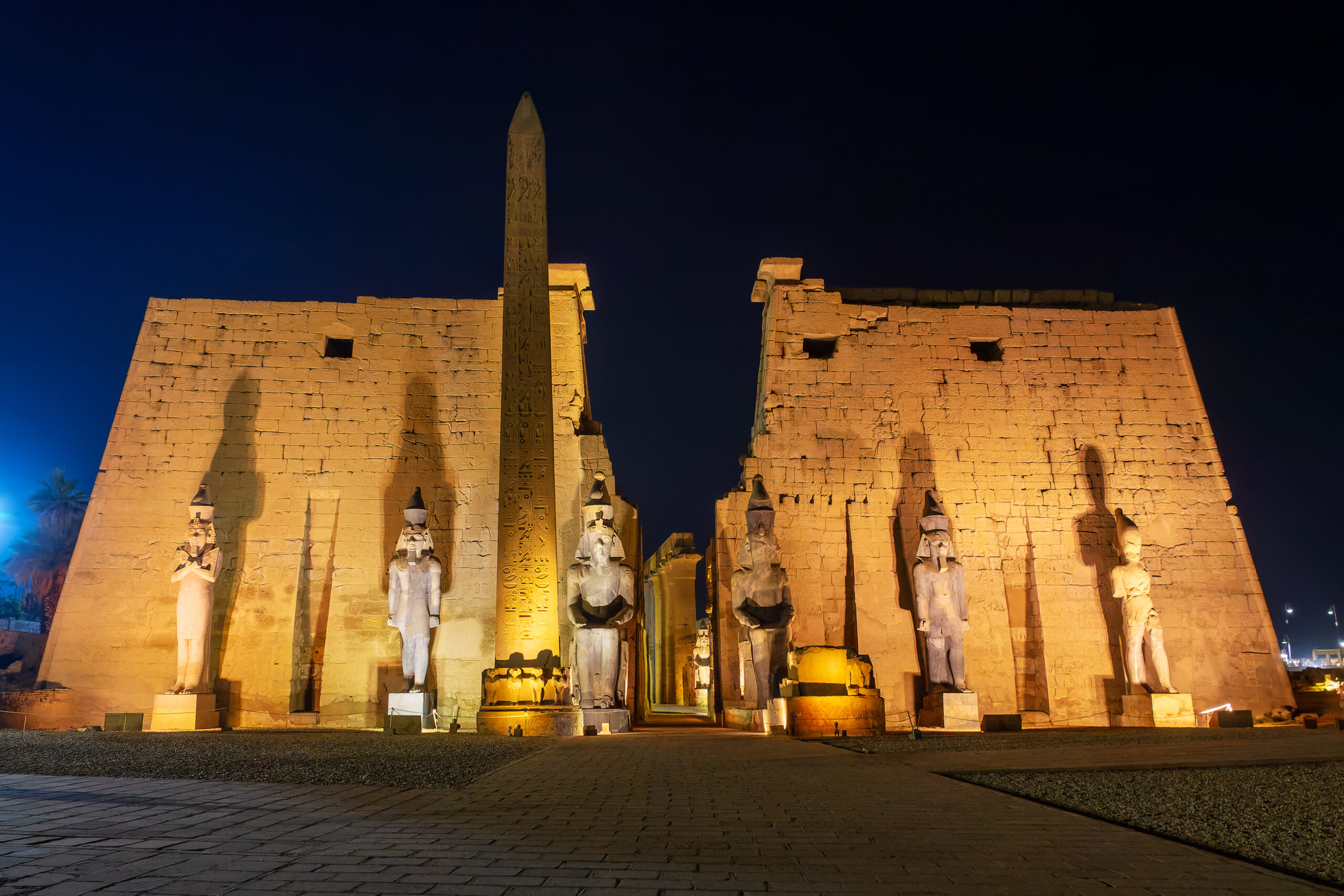 Ancient Egyptian temple facade with a tall hieroglyphic obelisk and statues, illuminated at night against a dark sky.