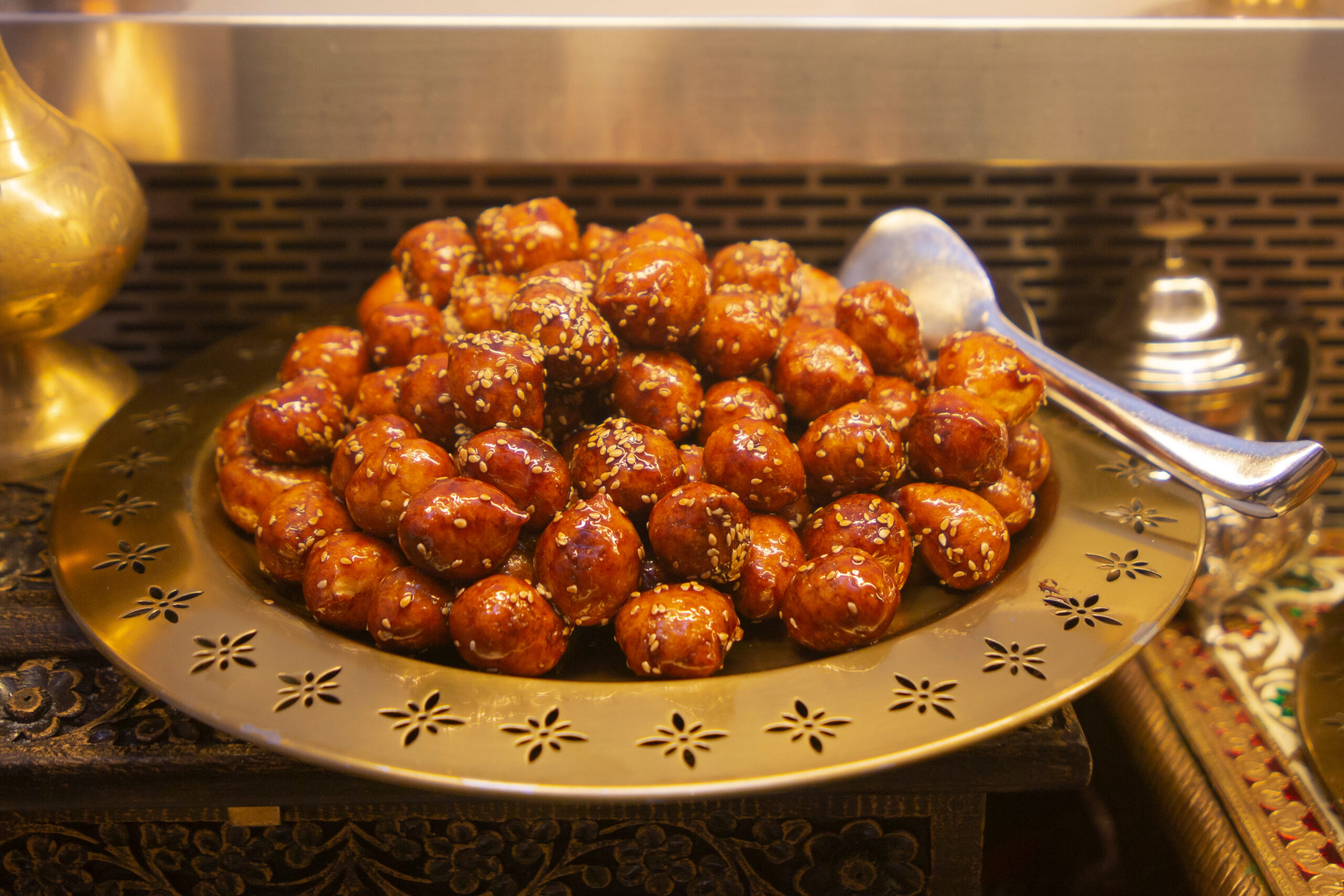 Pile of sesame-topped fried dough balls on a decorative metal platter at a buffet.