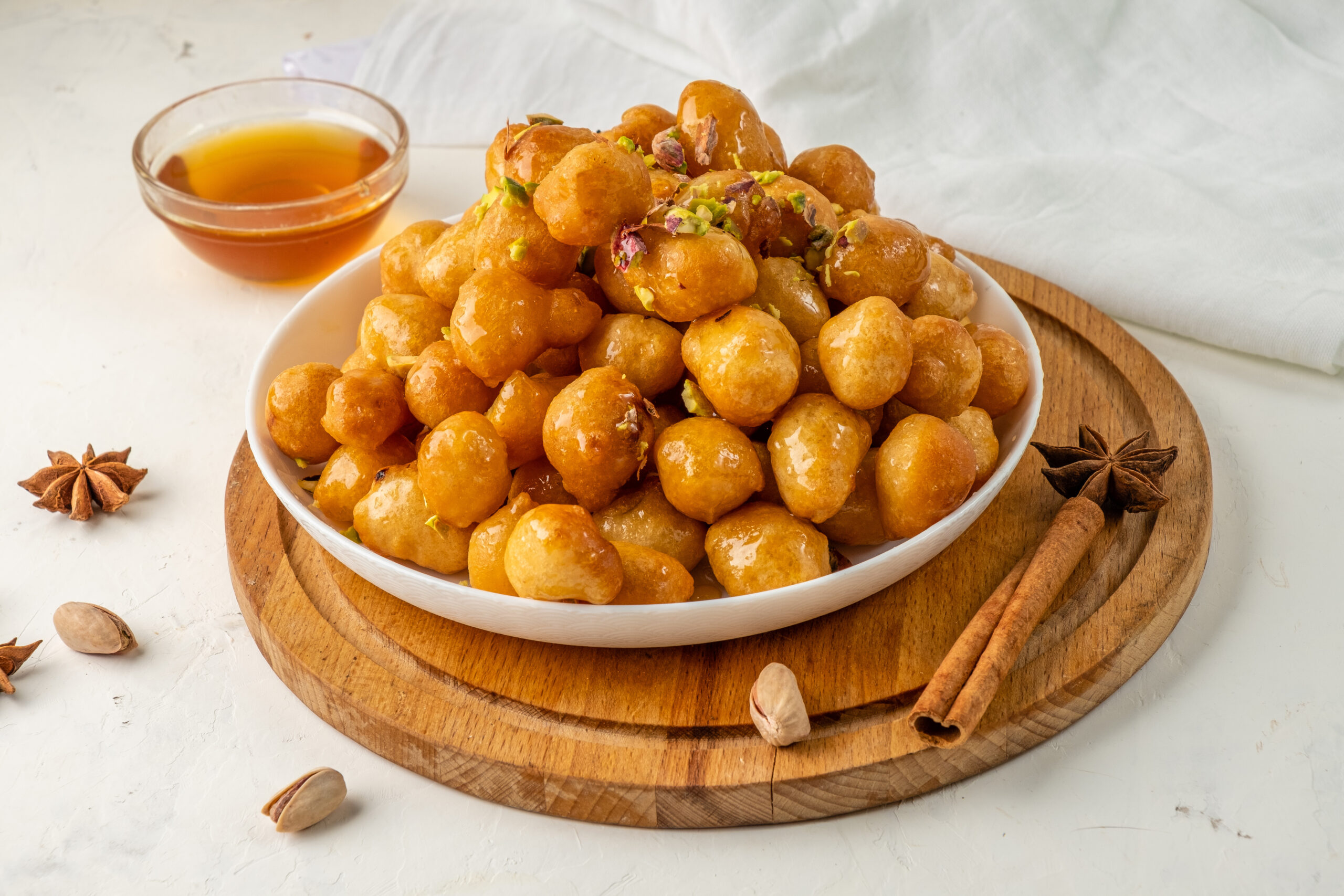 Bowl of syrup-soaked gulab jamun garnished with chopped pistachios on a wooden board with a small bowl of syrup nearby.