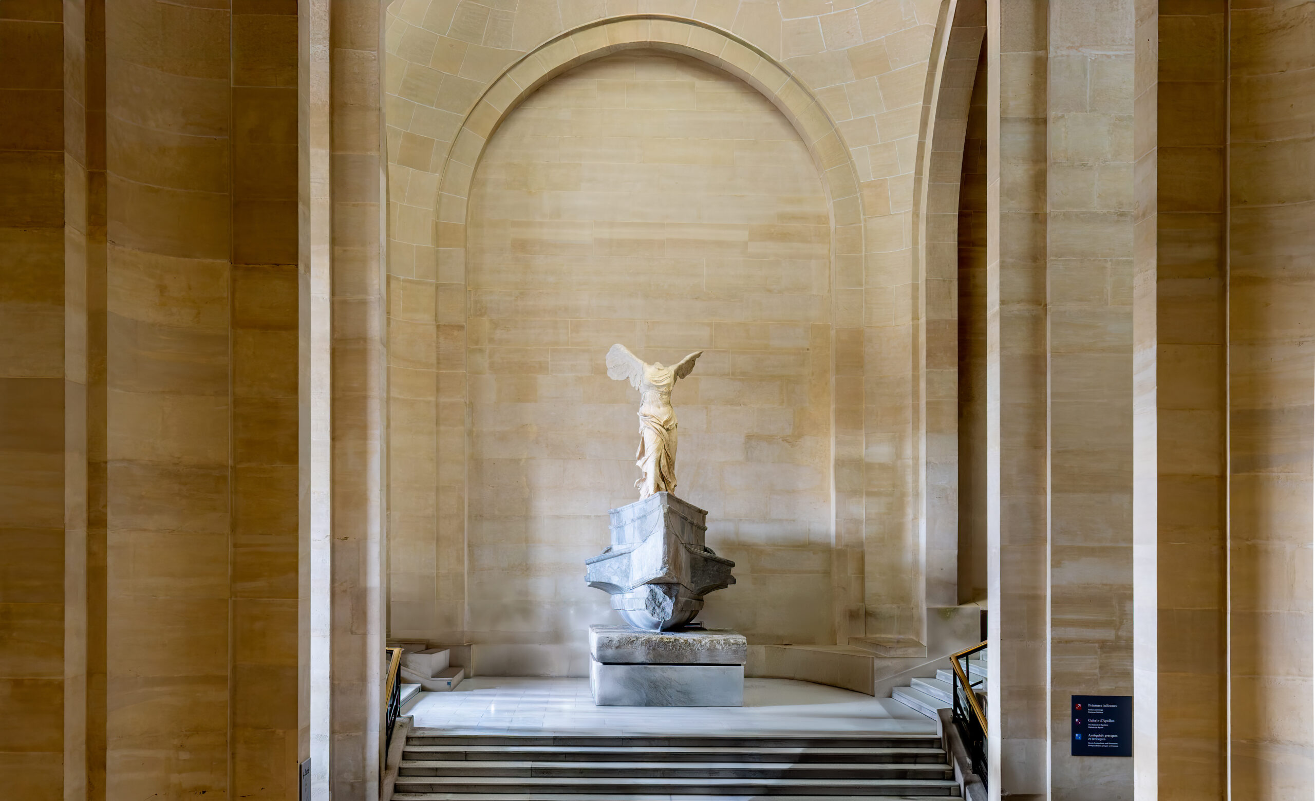 Statue of a winged figure on a pedestal in a grand sandstone hall with arches and steps behind it.