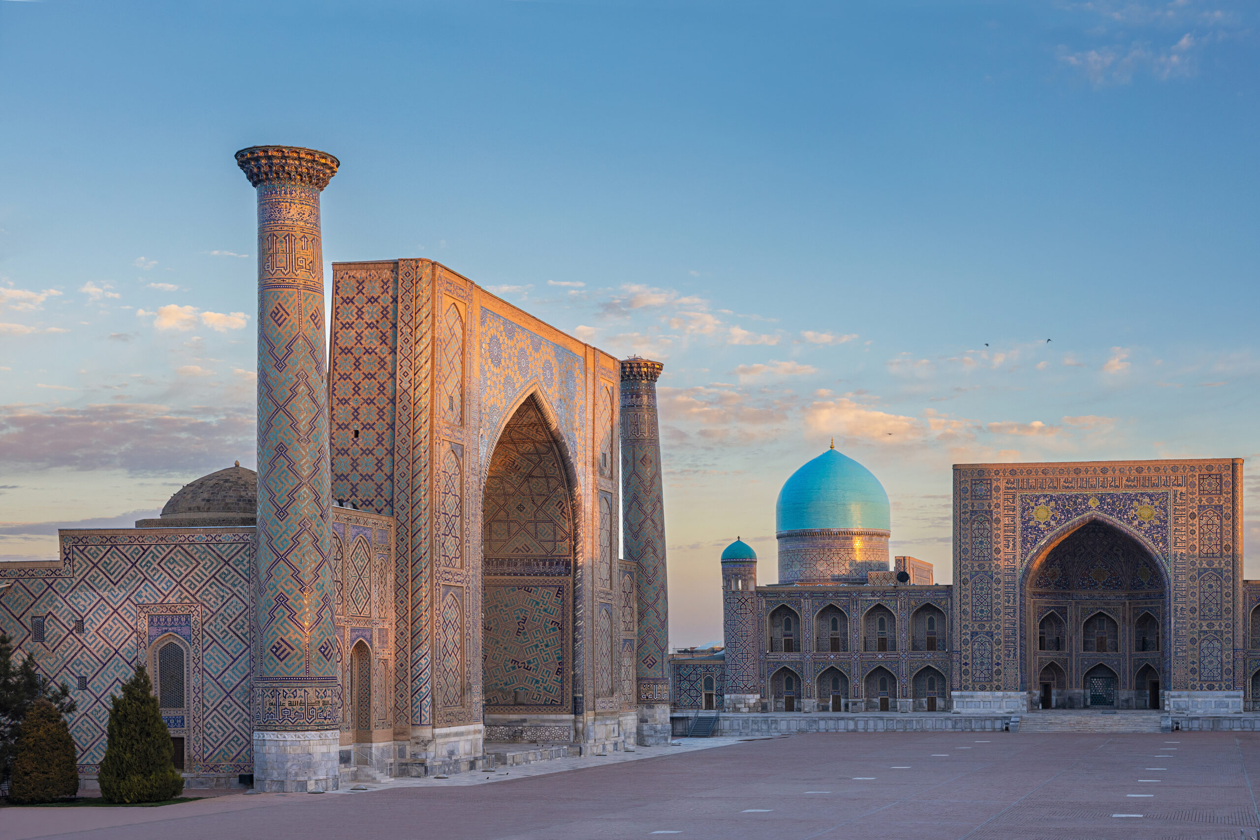 Ornate Islamic mosque complex with tall tiled minaret, blue domes, and arched gateways under a clear sky.