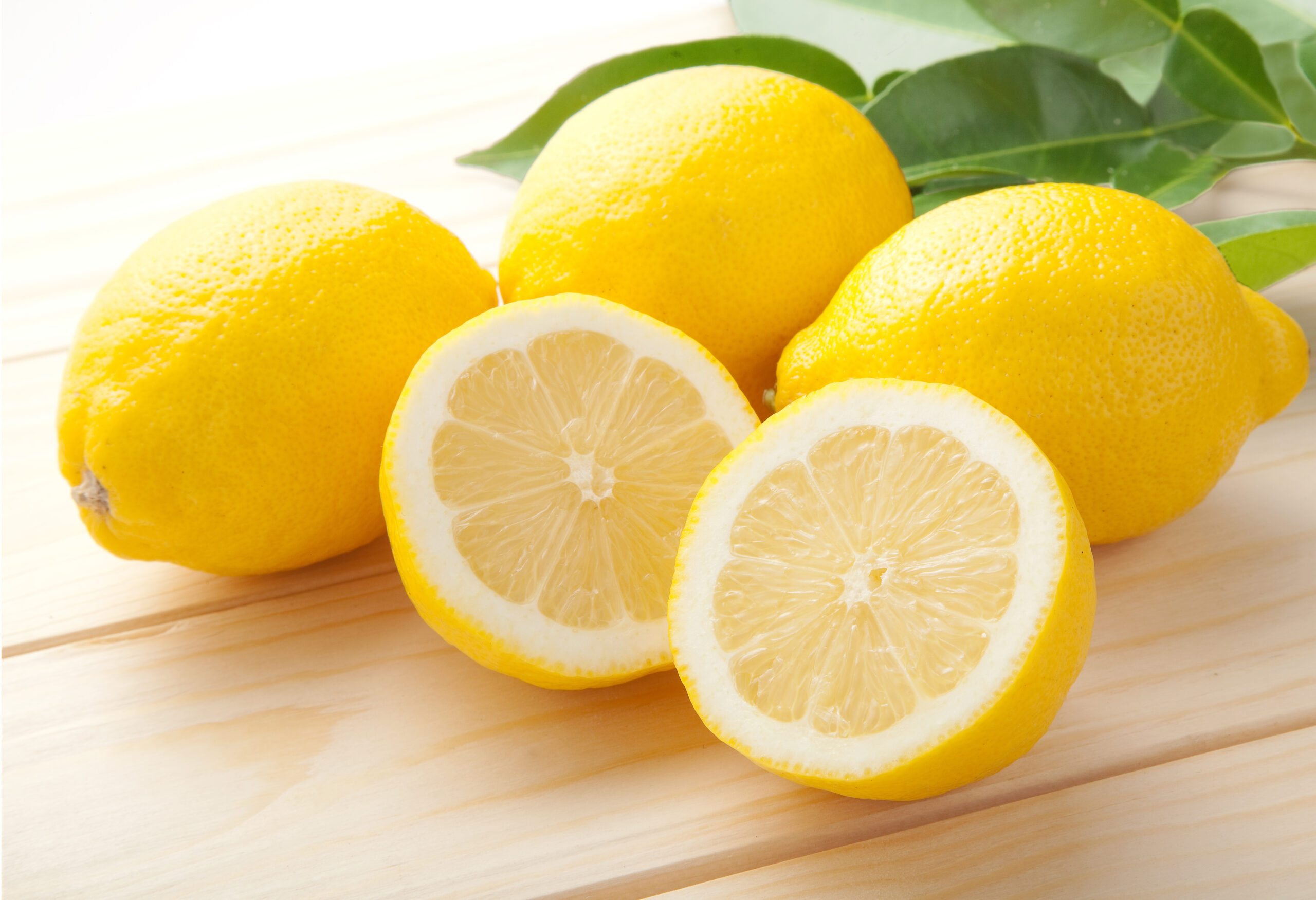 Bright yellow lemons with sliced halves on a light wooden table, with green leaves in the background