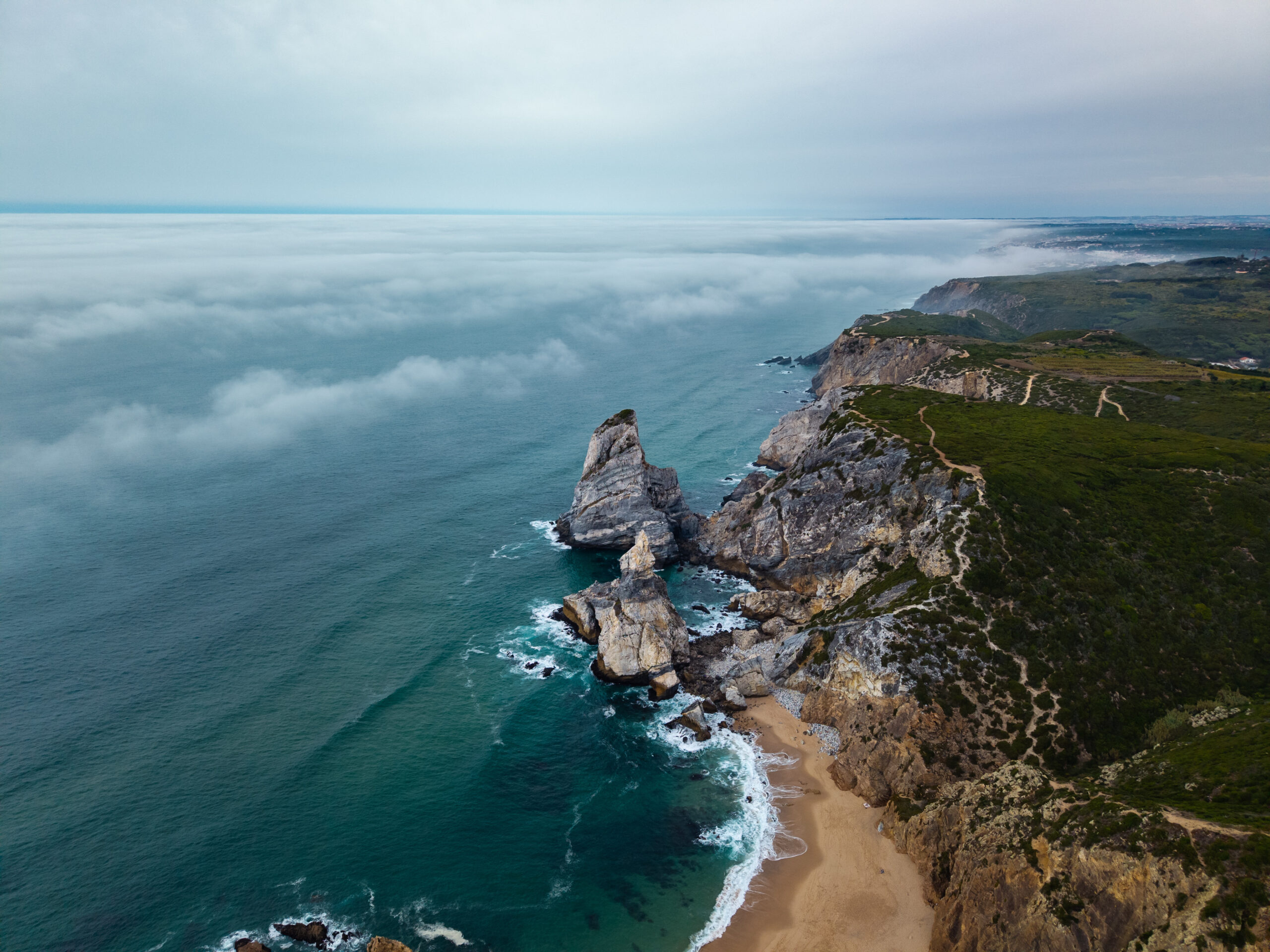 Aerial view of rugged coastline with steep cliffs, sandy beach below, and turquoise water on the left under a cloudy sky.