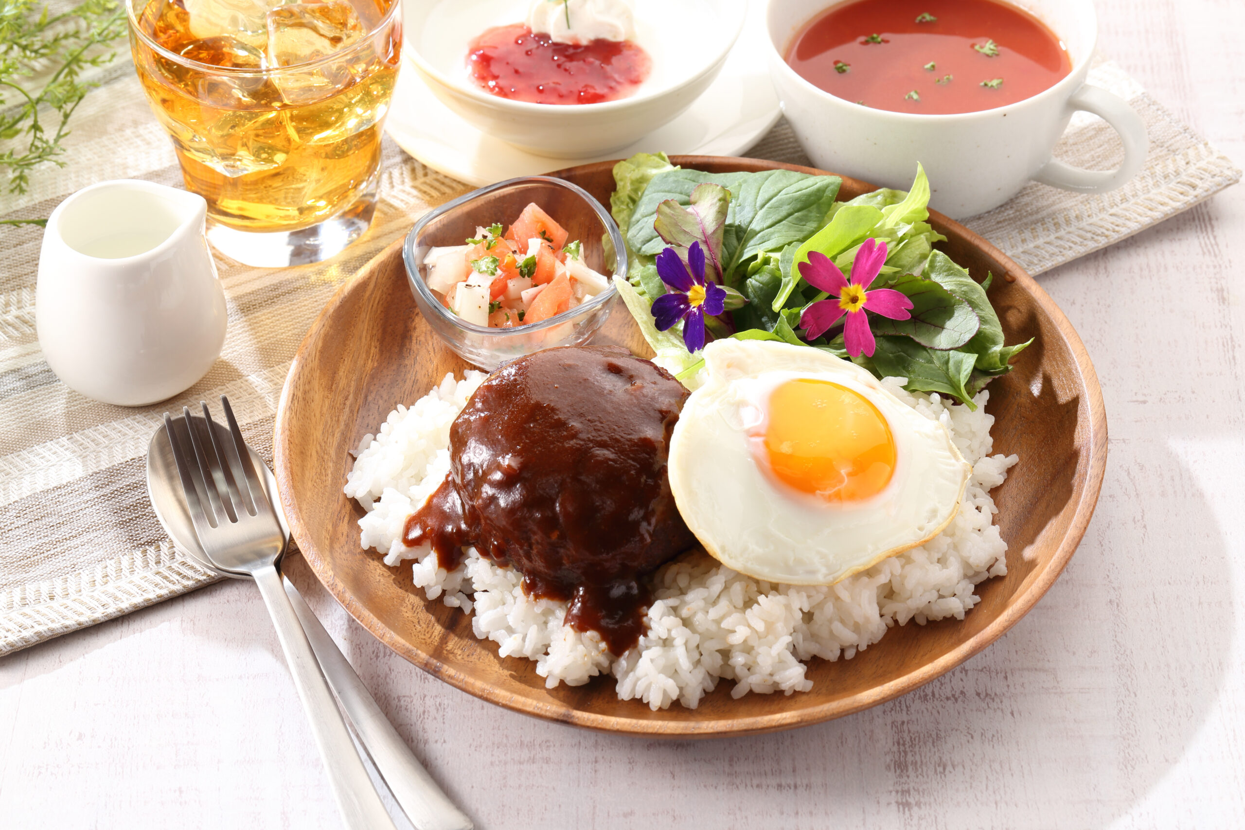 Plate of white rice with a glossy hamburger steak and sunny-side egg, plus greens, a small tomato salad, and colorful flowers on a wooden plate; beverages and soups in the background.