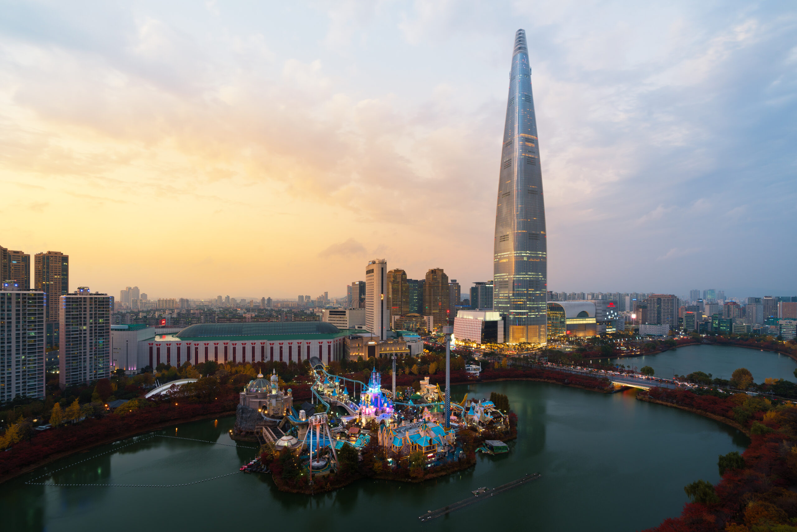 City skyline at sunset with the tall glass Bitexco Tower beside a river and a colorful amusement park in the foreground.