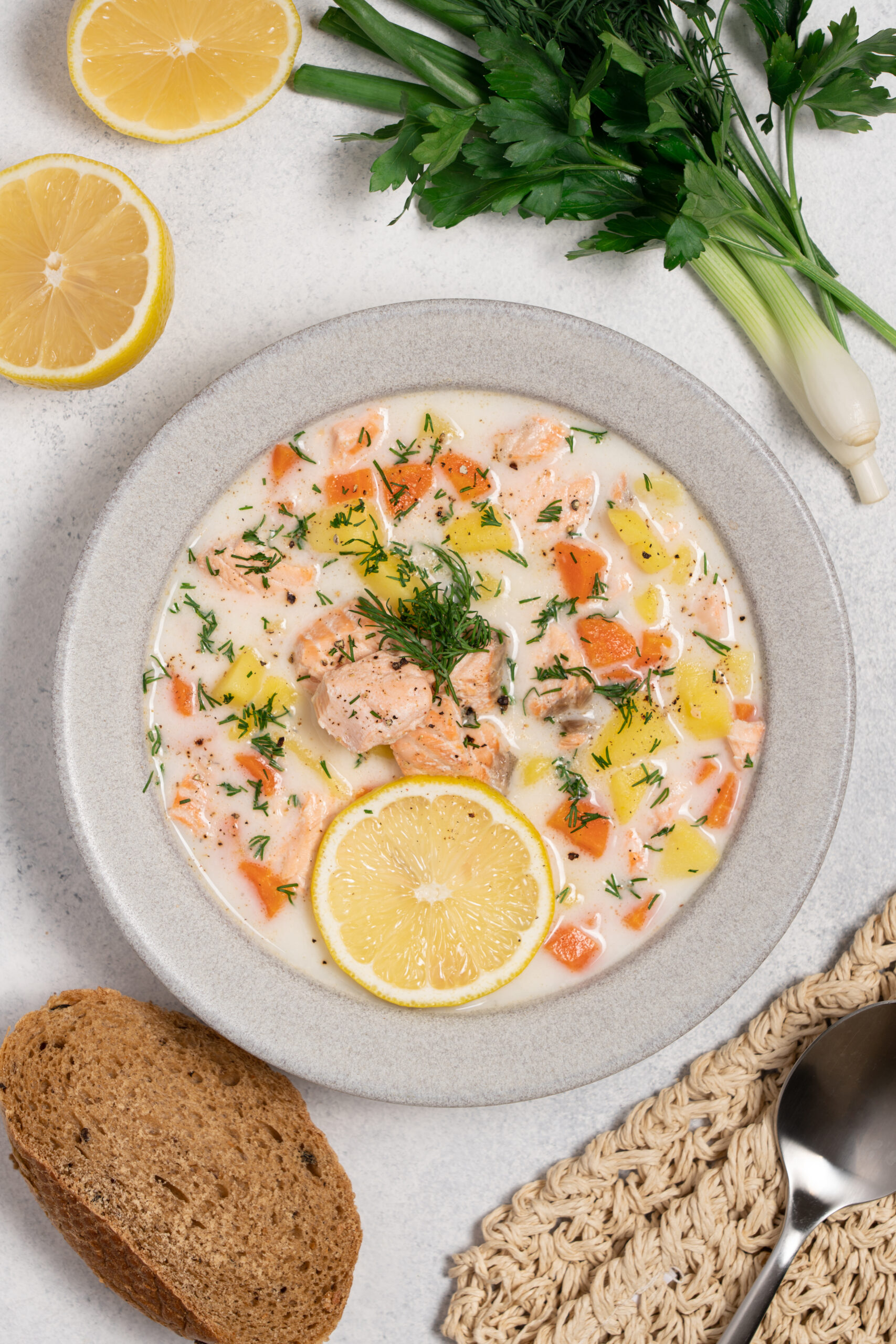Creamy salmon chowder with potatoes and carrots in a gray bowl, garnished with dill and lemon slices, with bread and a spoon nearby.