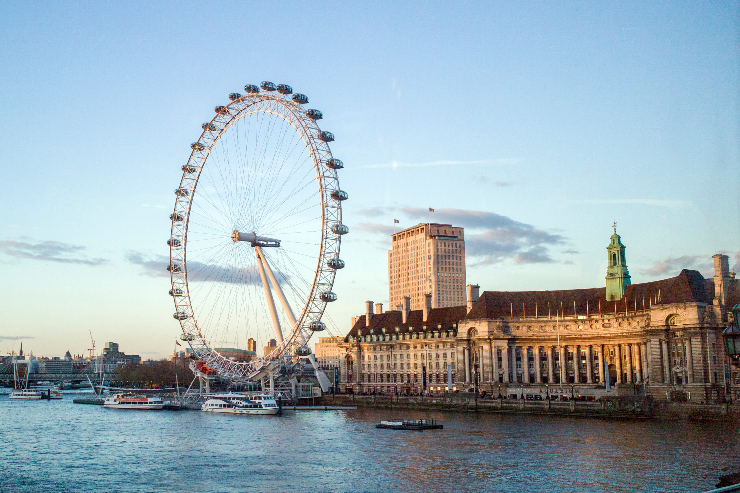 London Eye Ferris wheel along the River Thames with boats and historic buildings in the foreground under a blue sky.140 chars