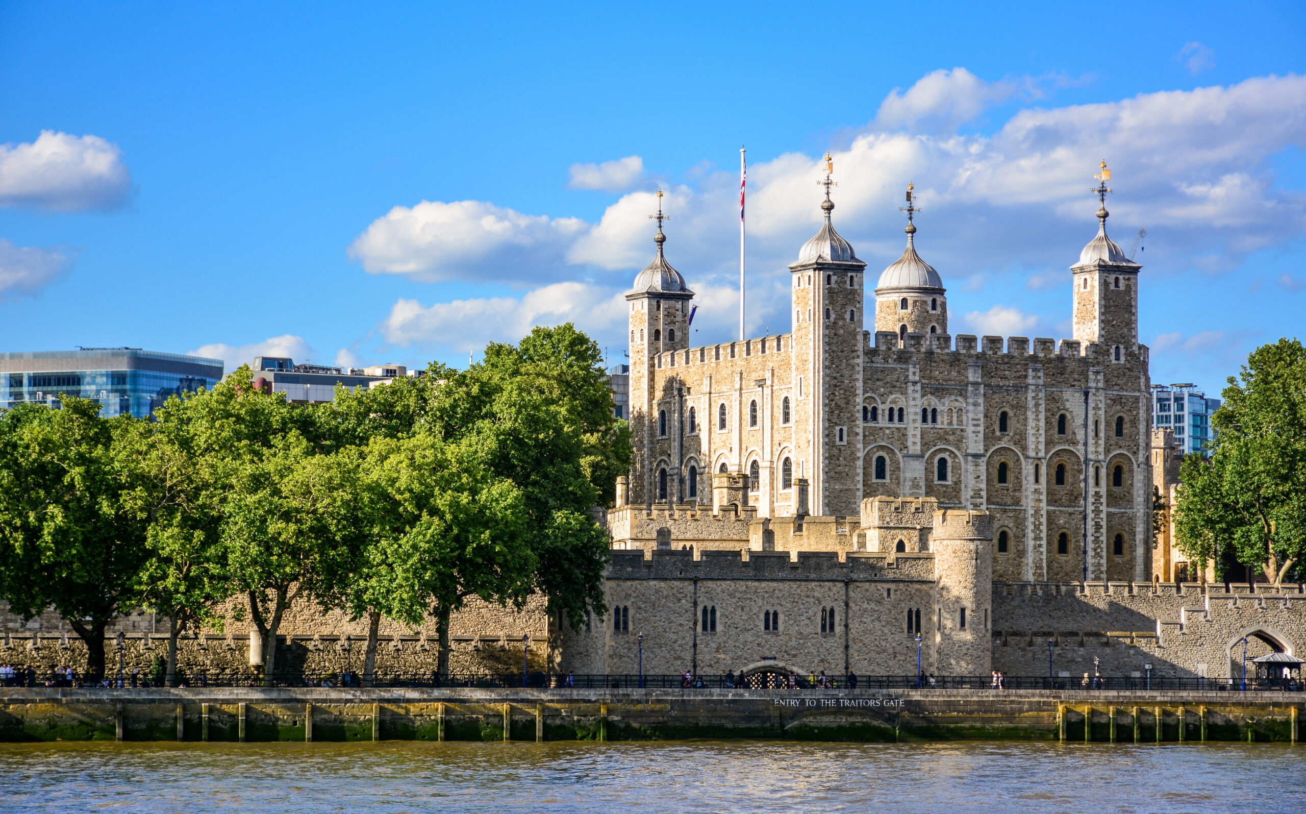 Tower of London along the River Thames with stone walls, multiple towers, and crenellations under a bright blue sky.