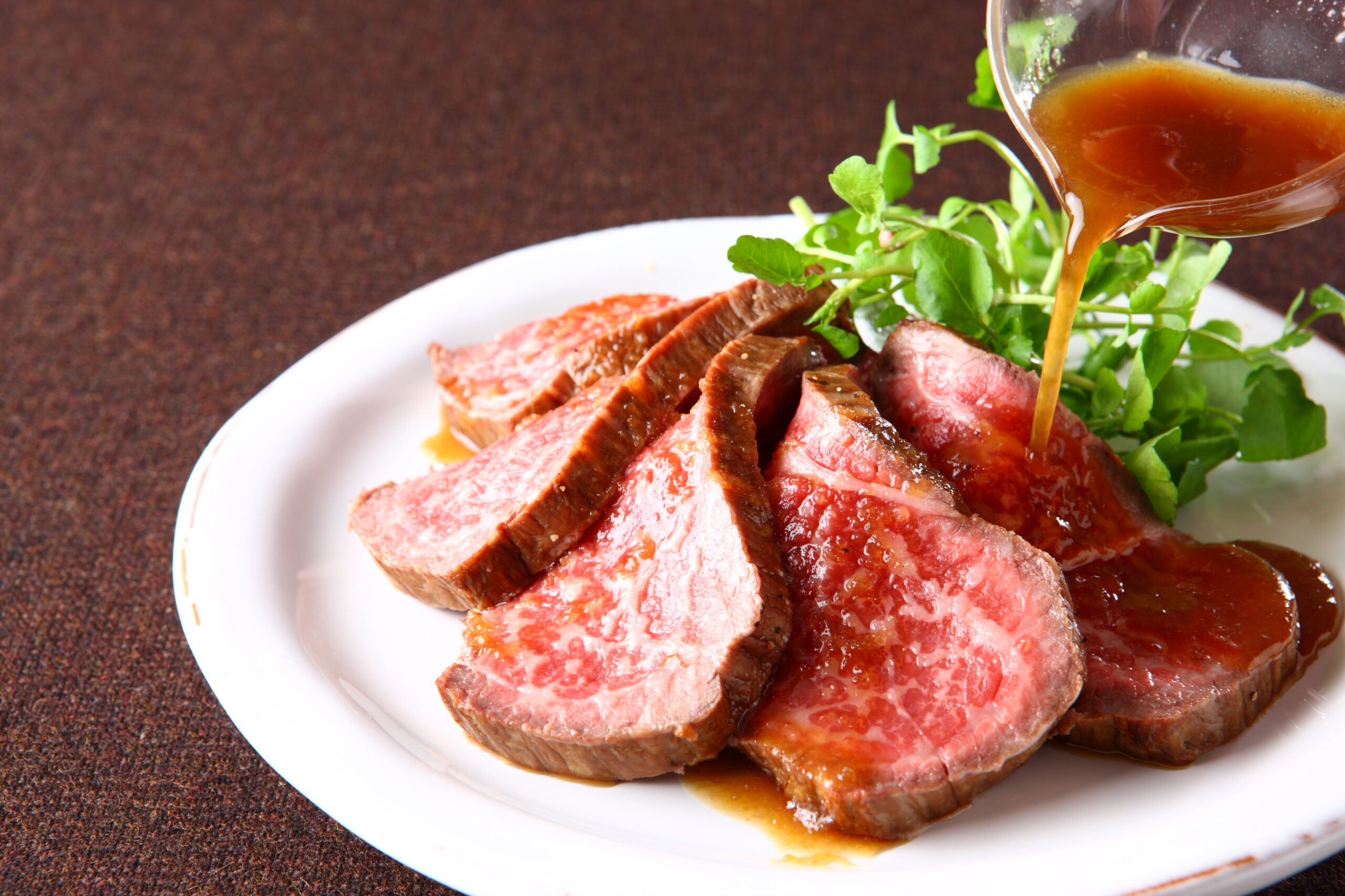 Slices of medium-rare steak on a white plate, with greens and sauce being poured over the meat.