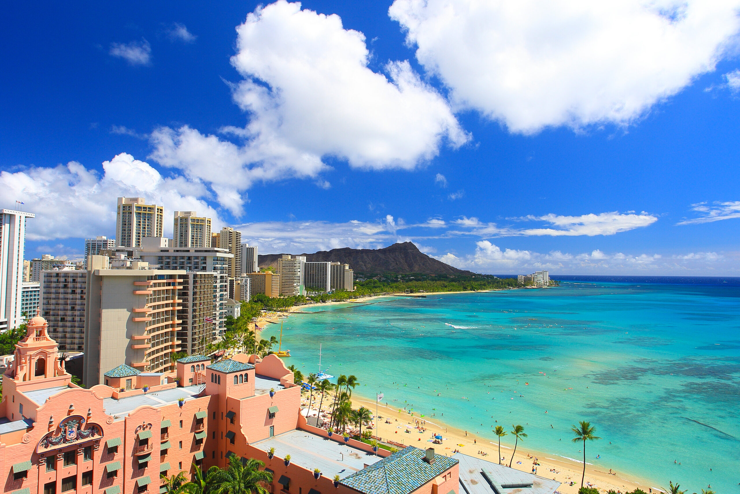 Coastal cityscape with pink Mediterranean-style buildings along a sandy beach and turquoise water under a bright blue sky with white clouds.