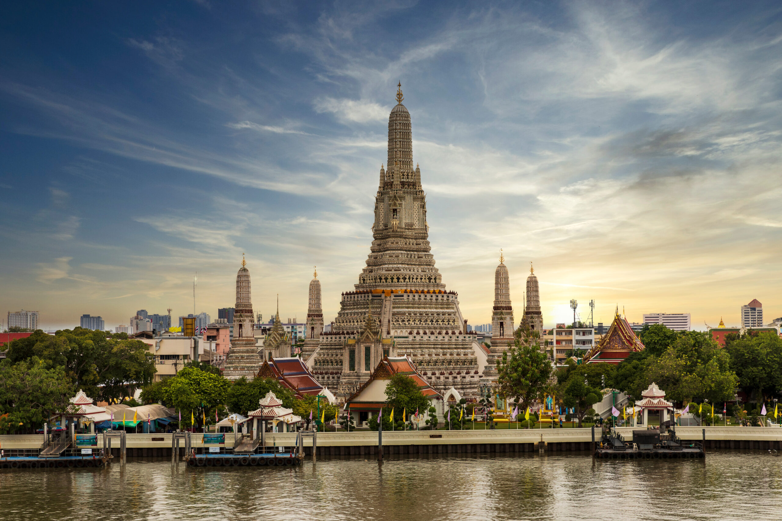 Wat Arun temple with a tall central prang and four surrounding towers along the river at sunset.
