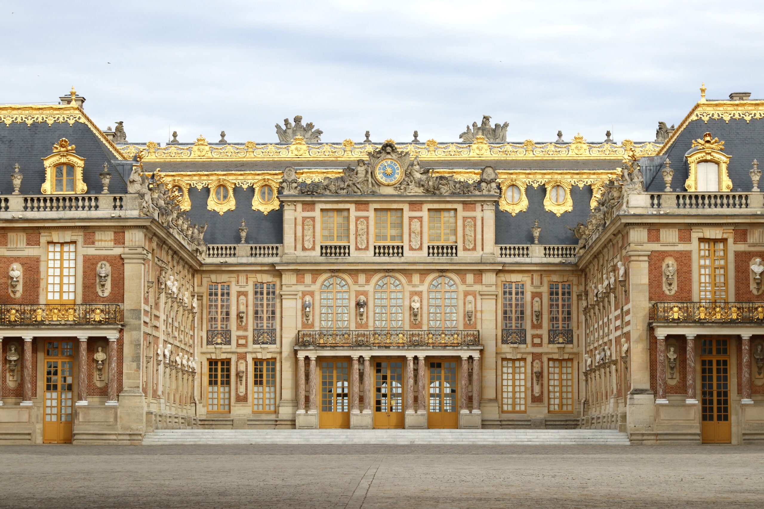Ornate palace facade with gilded decorations, statues, and a central clock, symmetrical wings on either side of yellow doors.