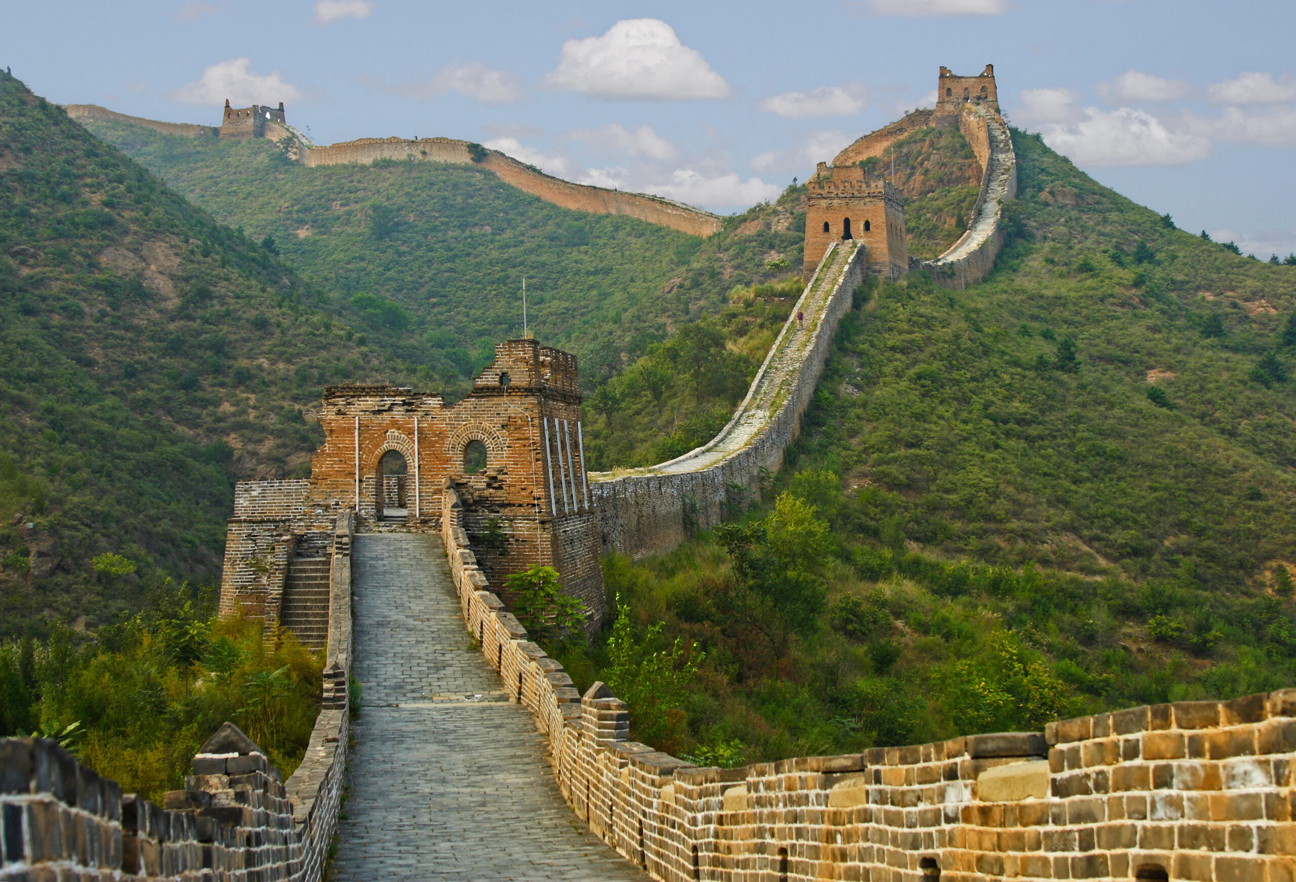 Section of the Great Wall winding over green hills beneath a blue sky with clouds, featuring watchtowers in the distance.