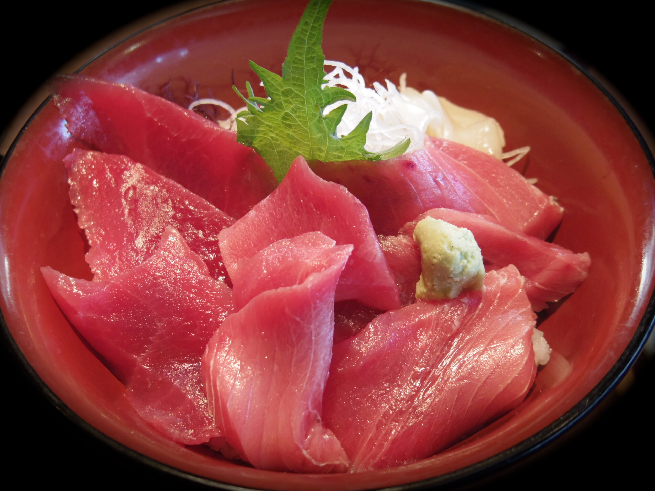 Sashimi bowl with thin tuna slices, a green shiso leaf, shredded daikon, and a dab of wasabi in a red bowl (edible dish).