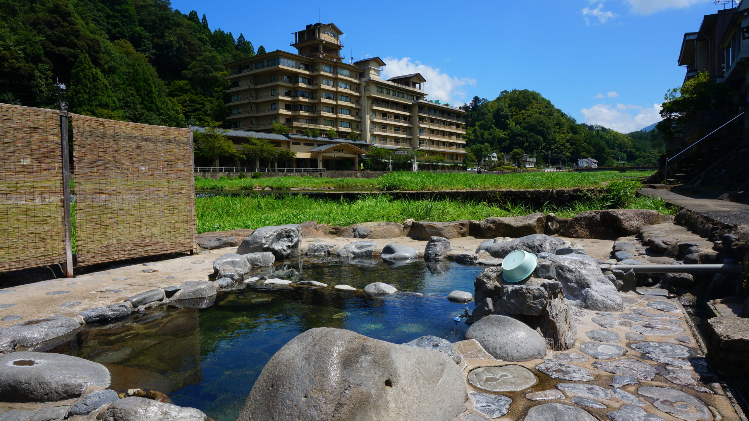 A riverside hotel with multiple balconies, set against green hills under a bright blue sky, with a rocky, shallow pool in the foreground and a bamboo screen on the left.