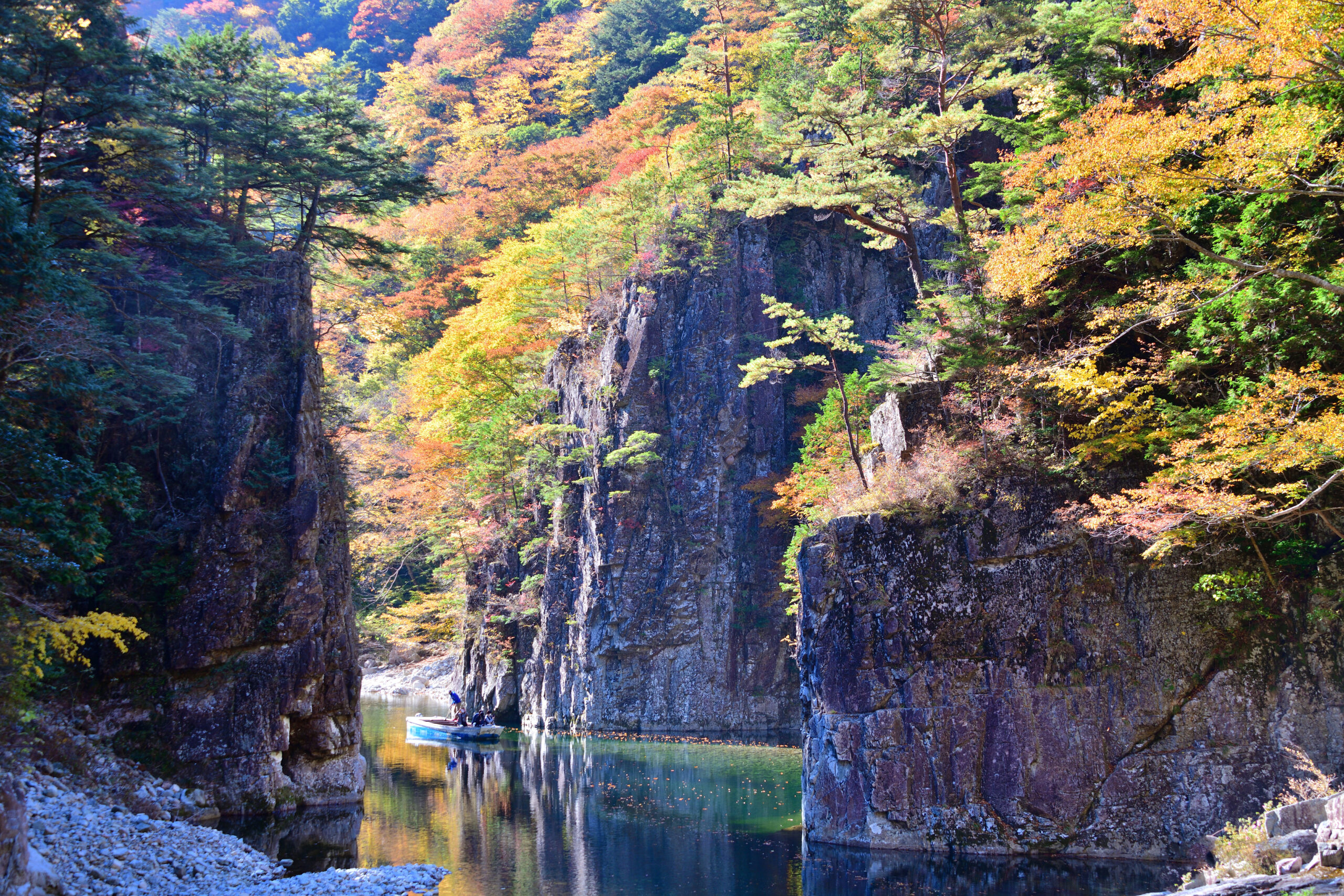 Autumn-colored trees line steep rocky canyon walls, with a calm river and a small boat carrying two people.