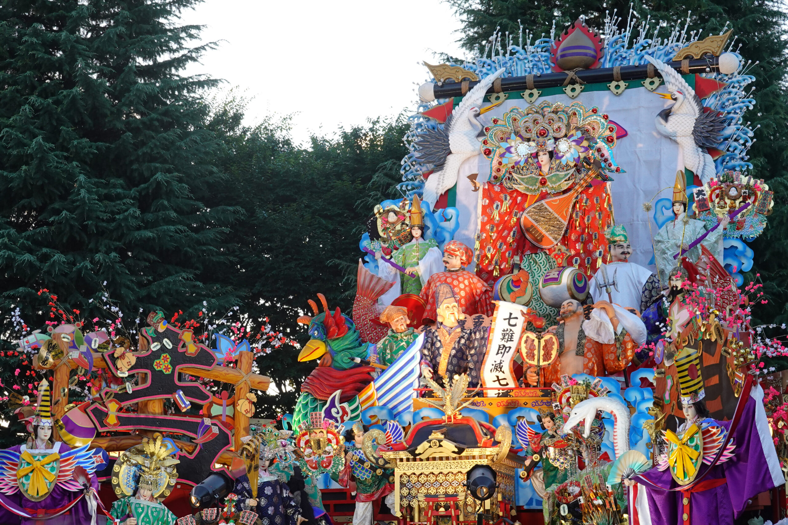 Colorful festival float with elaborately dressed Chinese opera figures, dragon and crane motifs, and ornate decorations against a green treeline.
