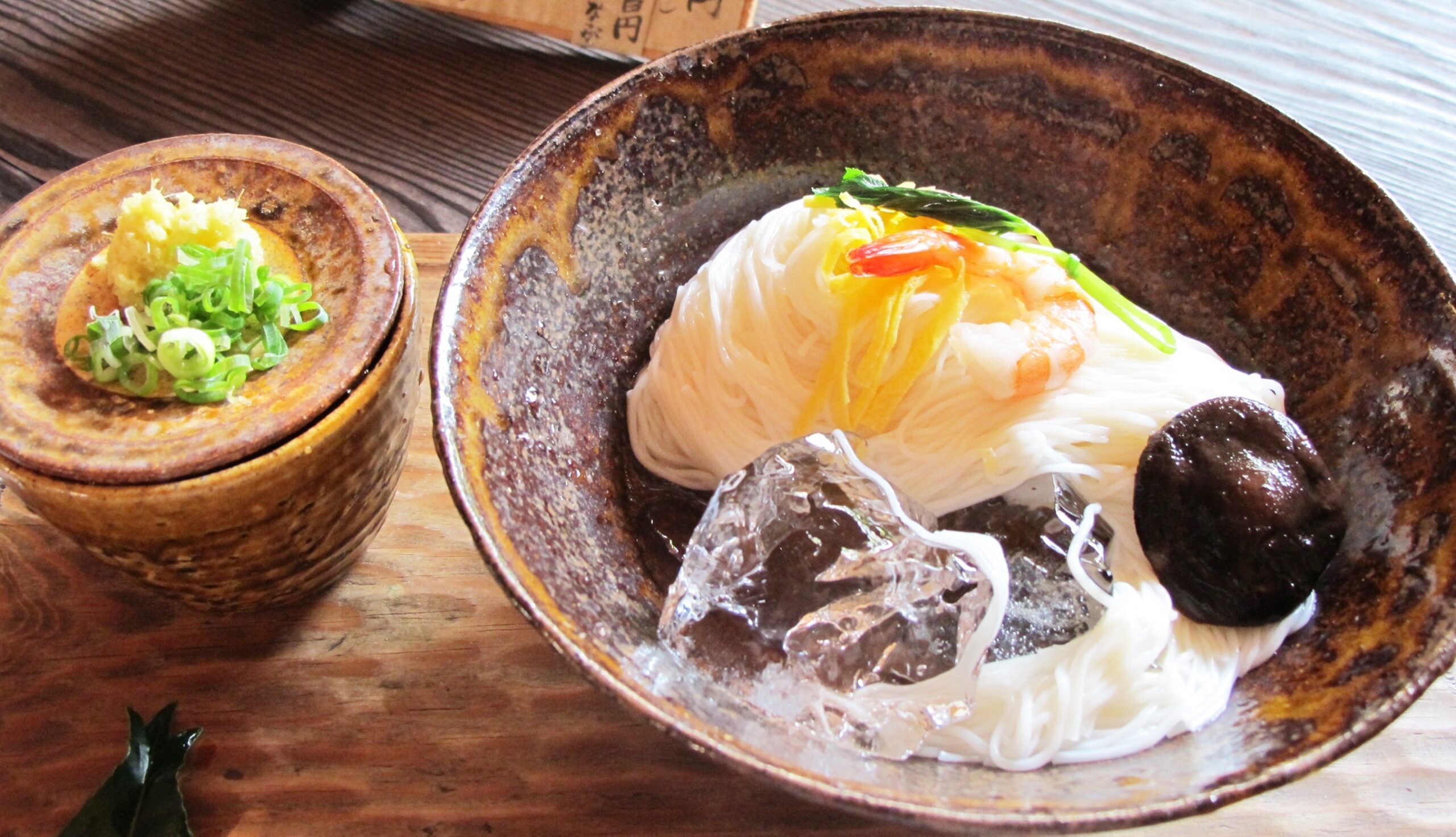 Rustic ceramic bowl with white thin noodles topped with yellow garnish and greens, beside a small bowl of grated ginger and chopped scallions on a wooden table