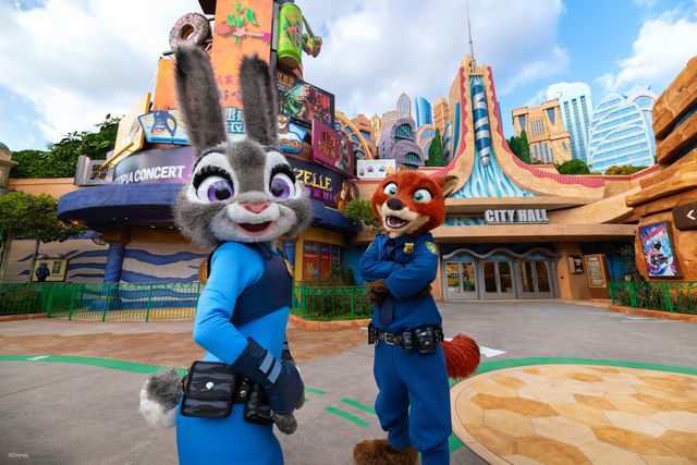 Two costumed characters—a gray rabbit and a red fox—pose for the camera outside City Hall in a colorful theme park.