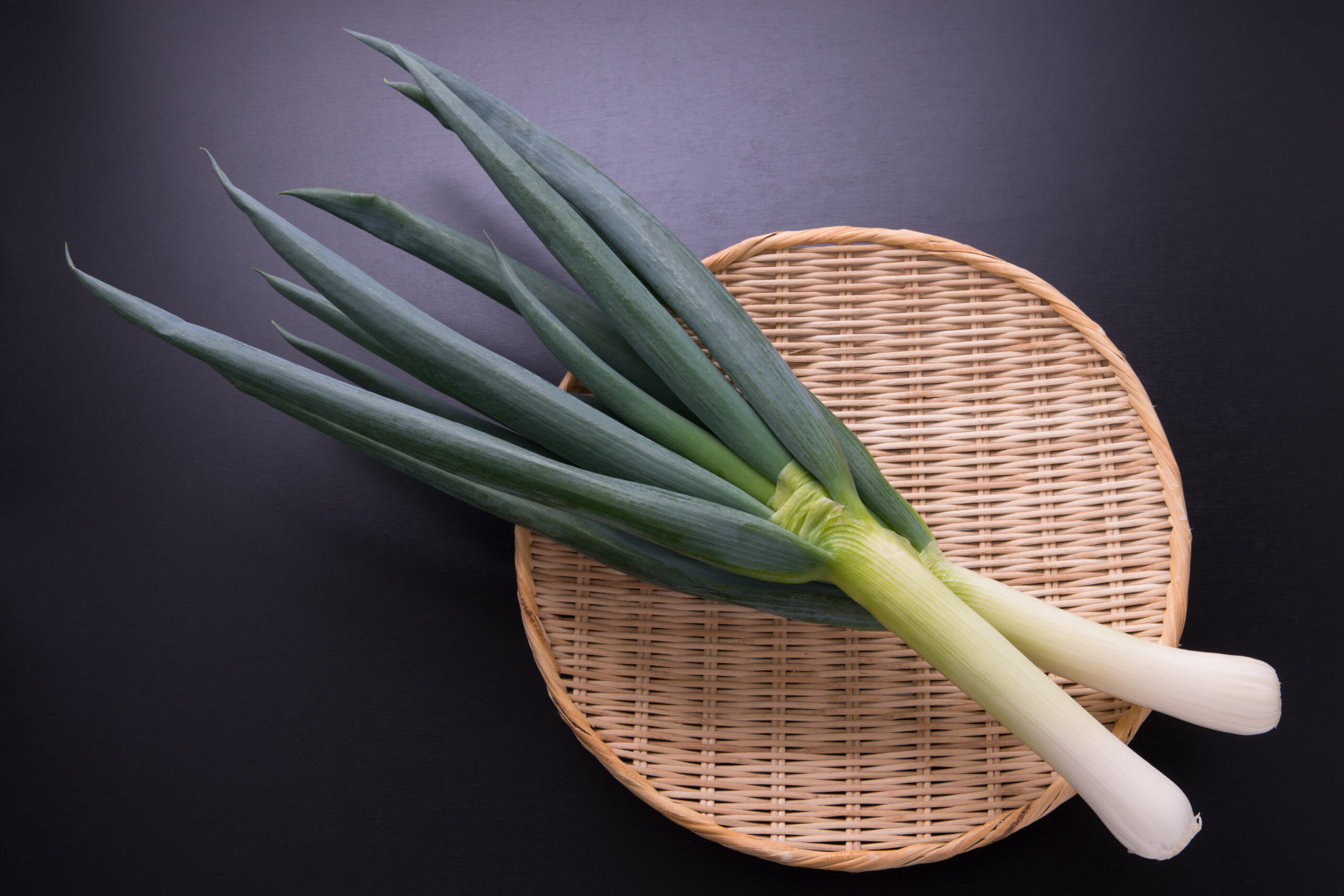 Bunch of green leeks with white stalks laid in a round wicker basket on a dark background.