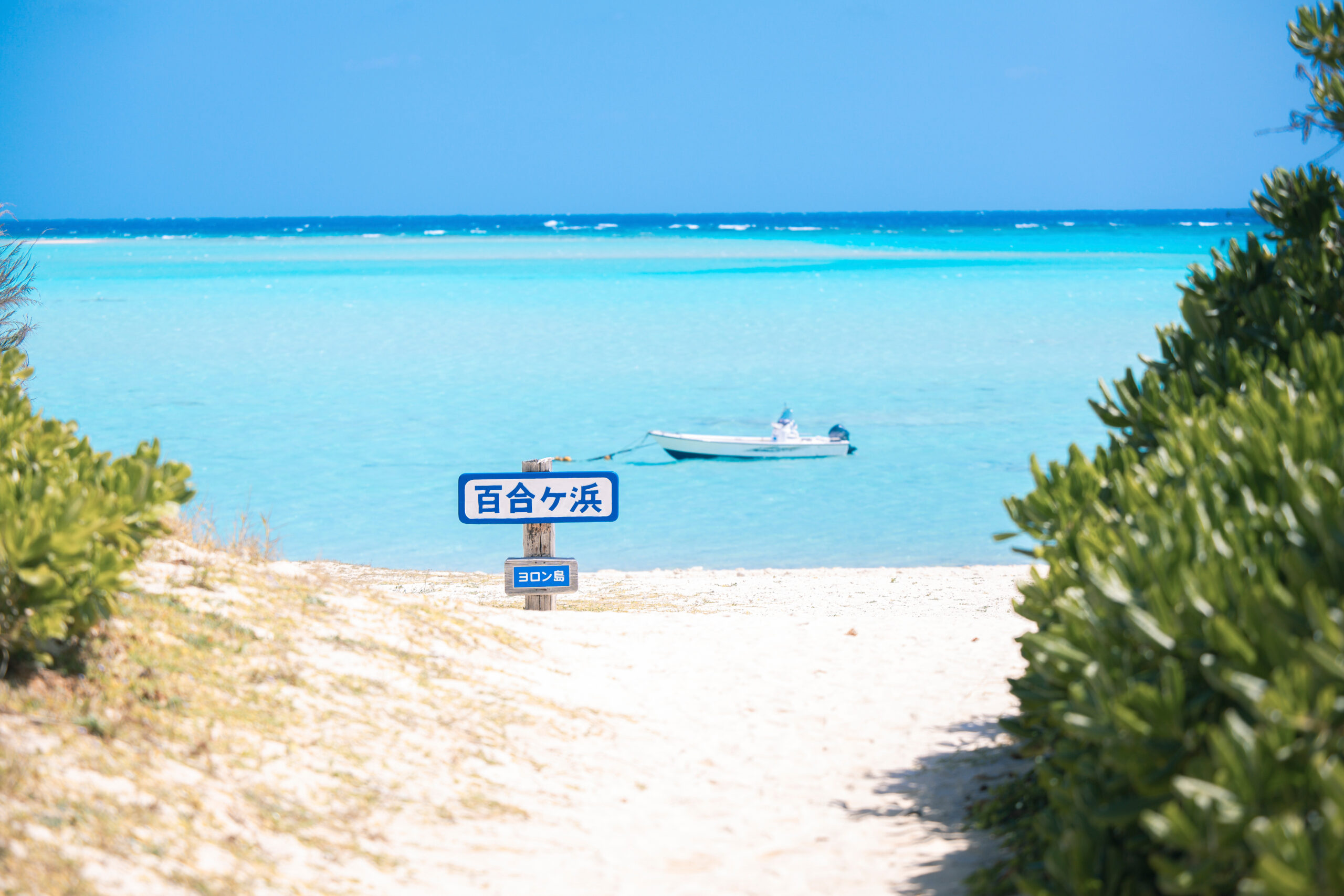 Beach scene with turquoise water, small boat in the bay, and a sign in Japanese in the foreground reading 百合ヶ浜 and ヨロン島 (Yoron Island).