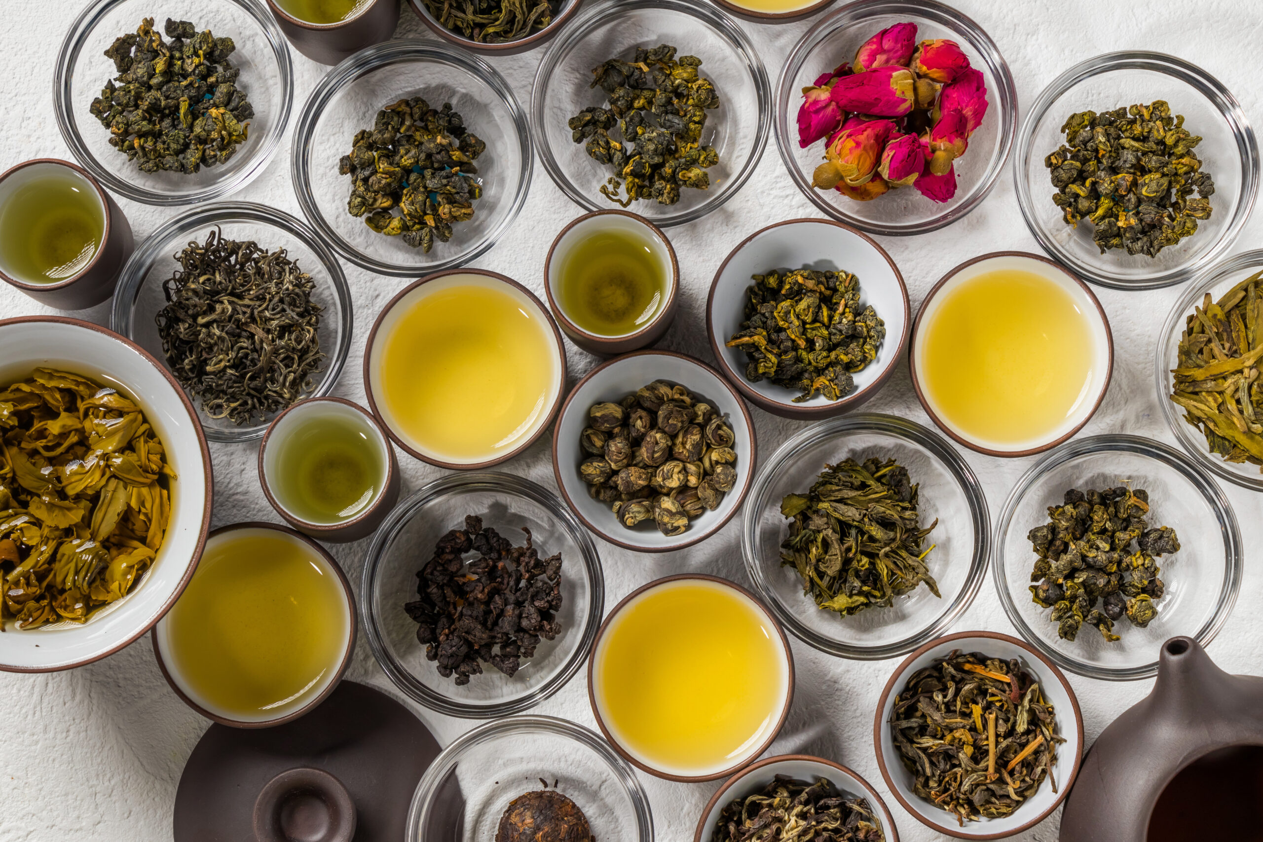 Assortment of loose tea leaves in glass bowls with several cups of pale yellow tea on a white surface.