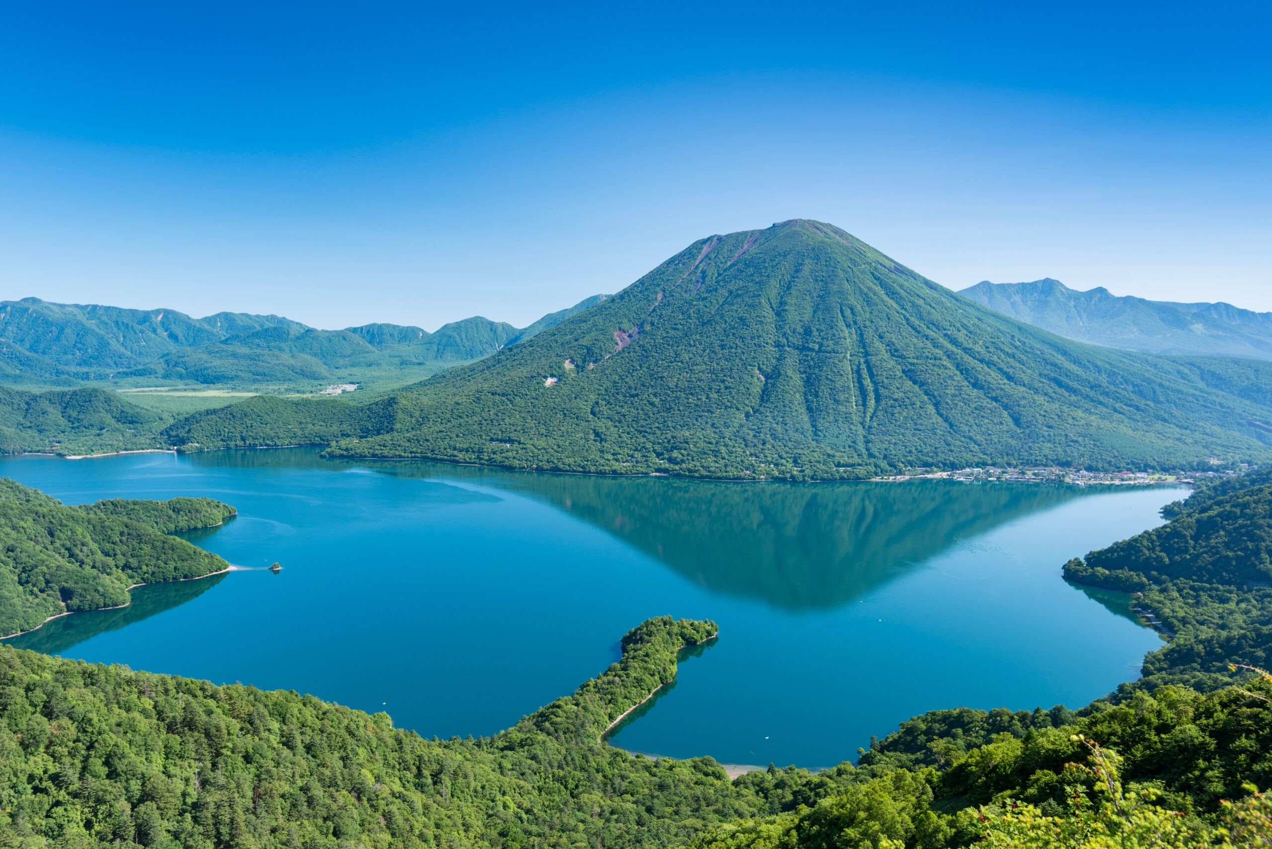 Aerial view of a forested mountain beside a blue lake under a clear blue sky in a wide landscape.