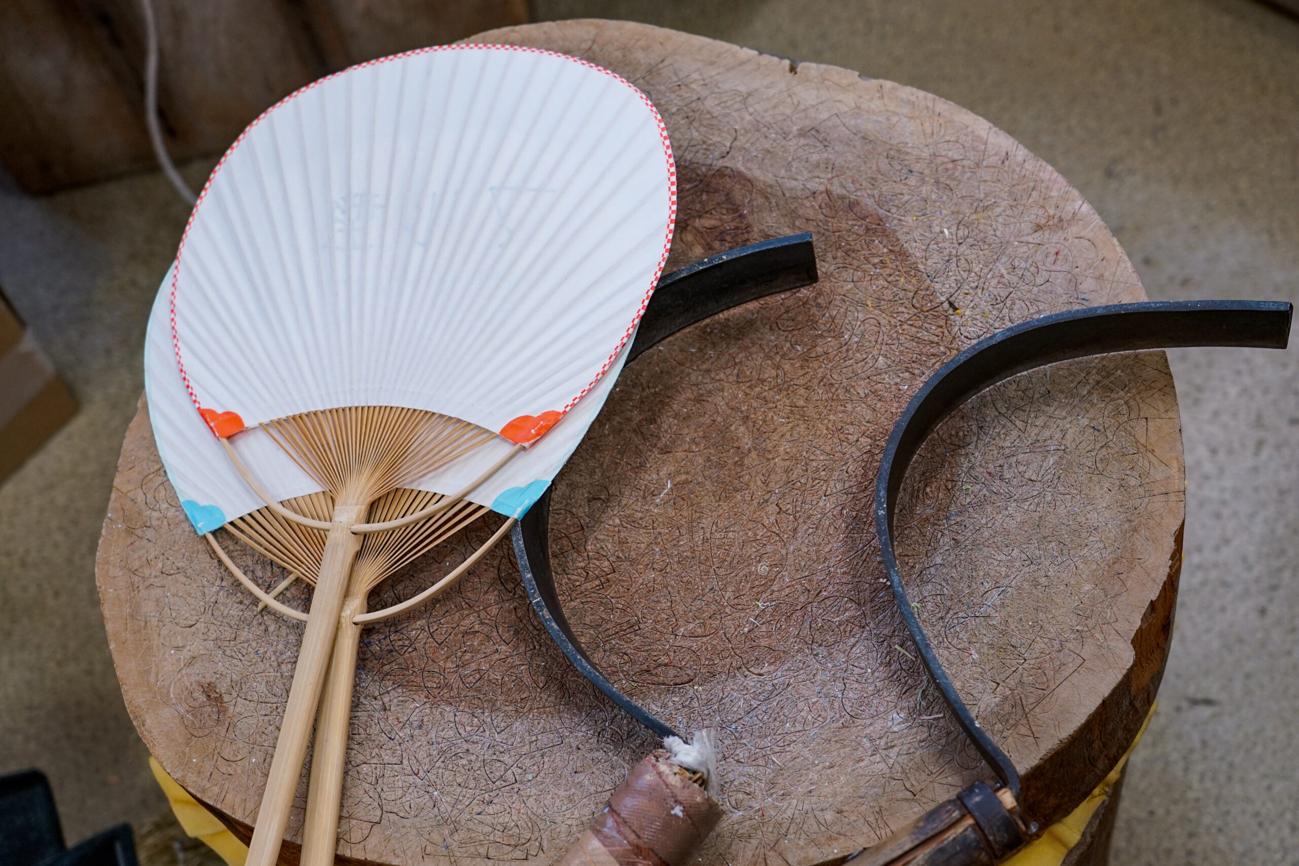 Traditional white paper handheld fan with wooden ribs resting on a weathered round wooden table in a workshop setting.