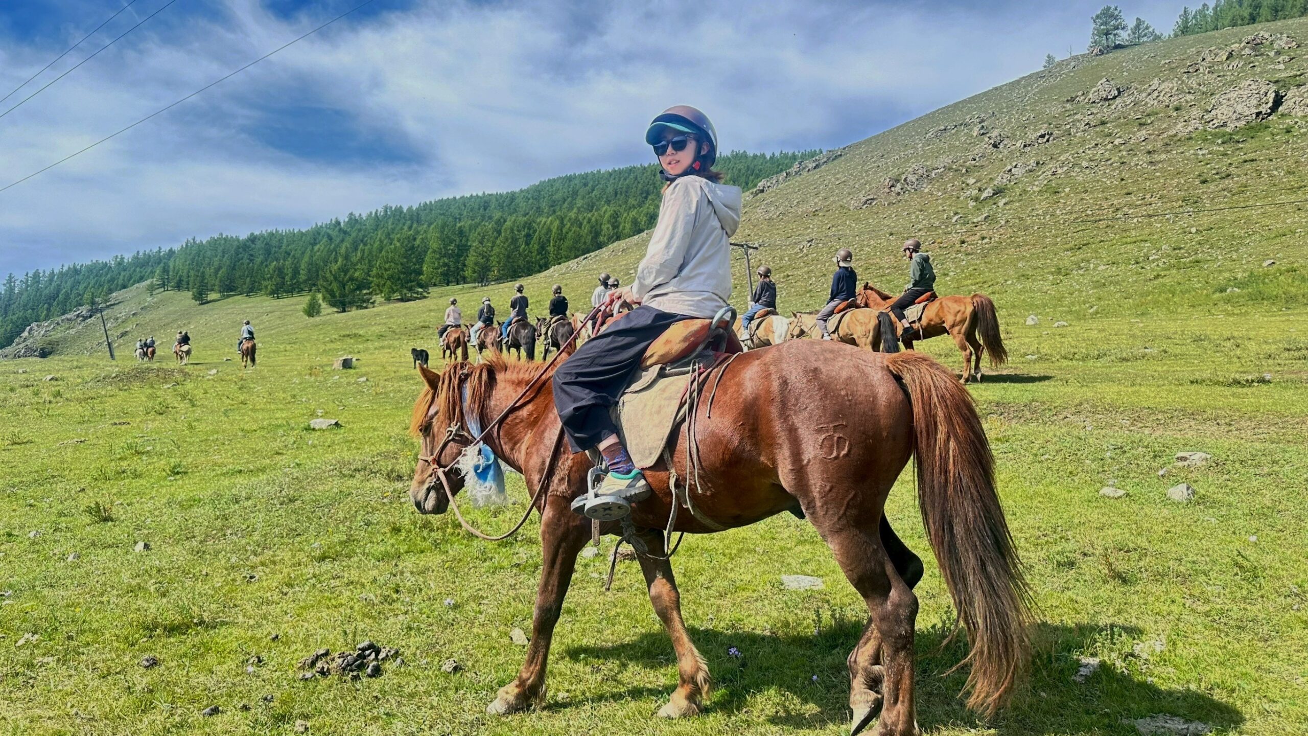 Group of riders on horseback crossing a grassy hillside, with a forested slope in the background and a rider in the foreground looking toward the camera.