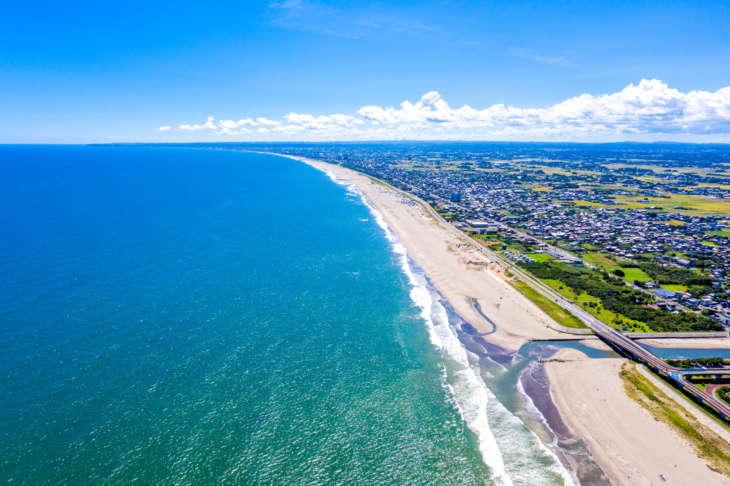 Aerial view of a curved sandy beach along a blue ocean with a town and green fields inland.