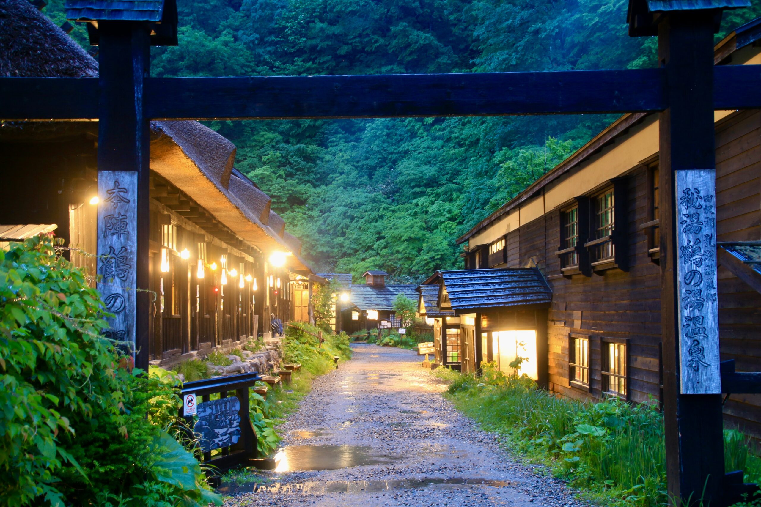 Narrow wooden street between traditional Japanese buildings with warm lanterns along a gravel path and green forested hills in the background.