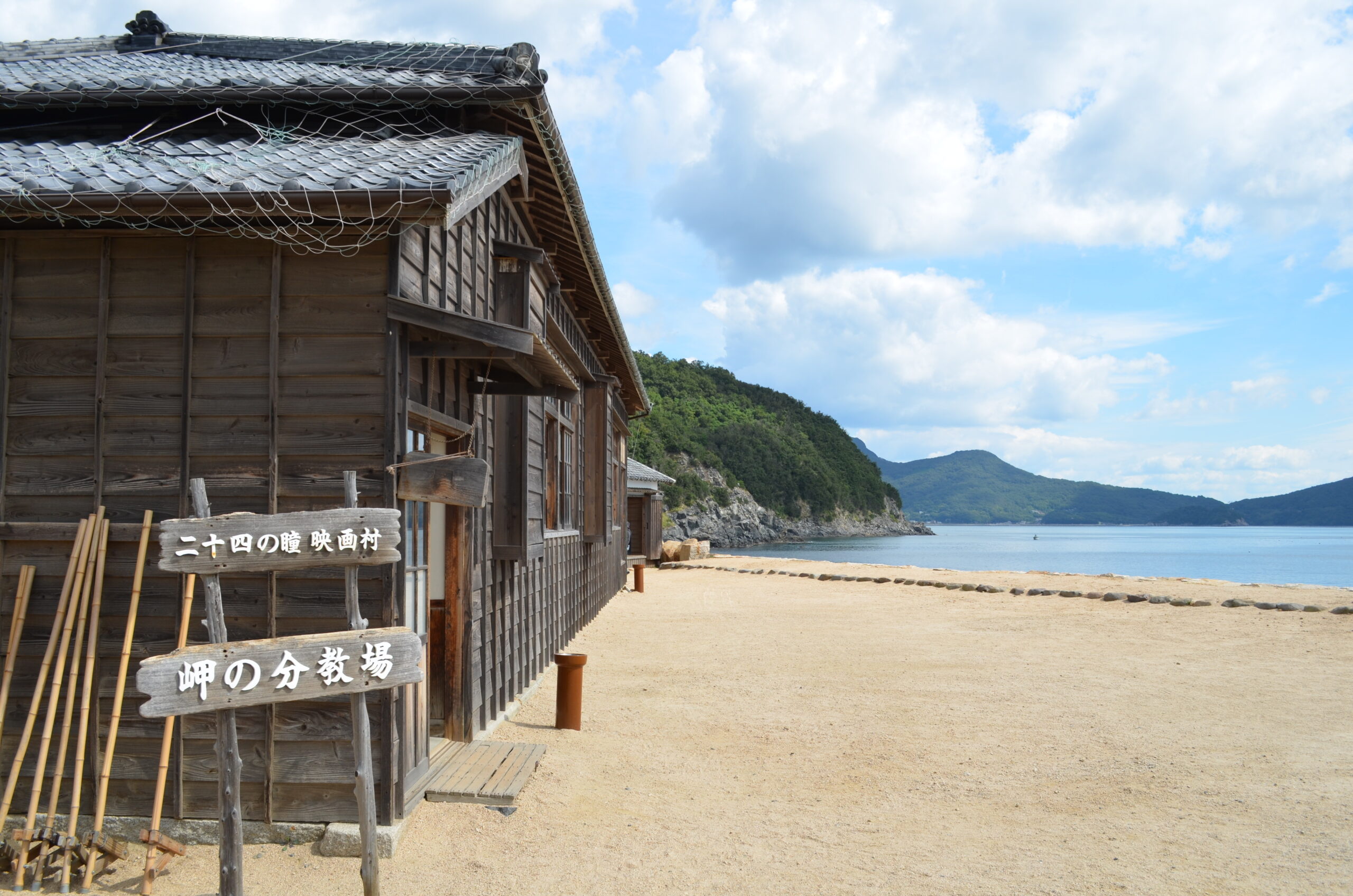 Weathered wooden coastal building with fishing nets on the roof, bamboo poles outside, and a sandy beach with blue sea beyond.