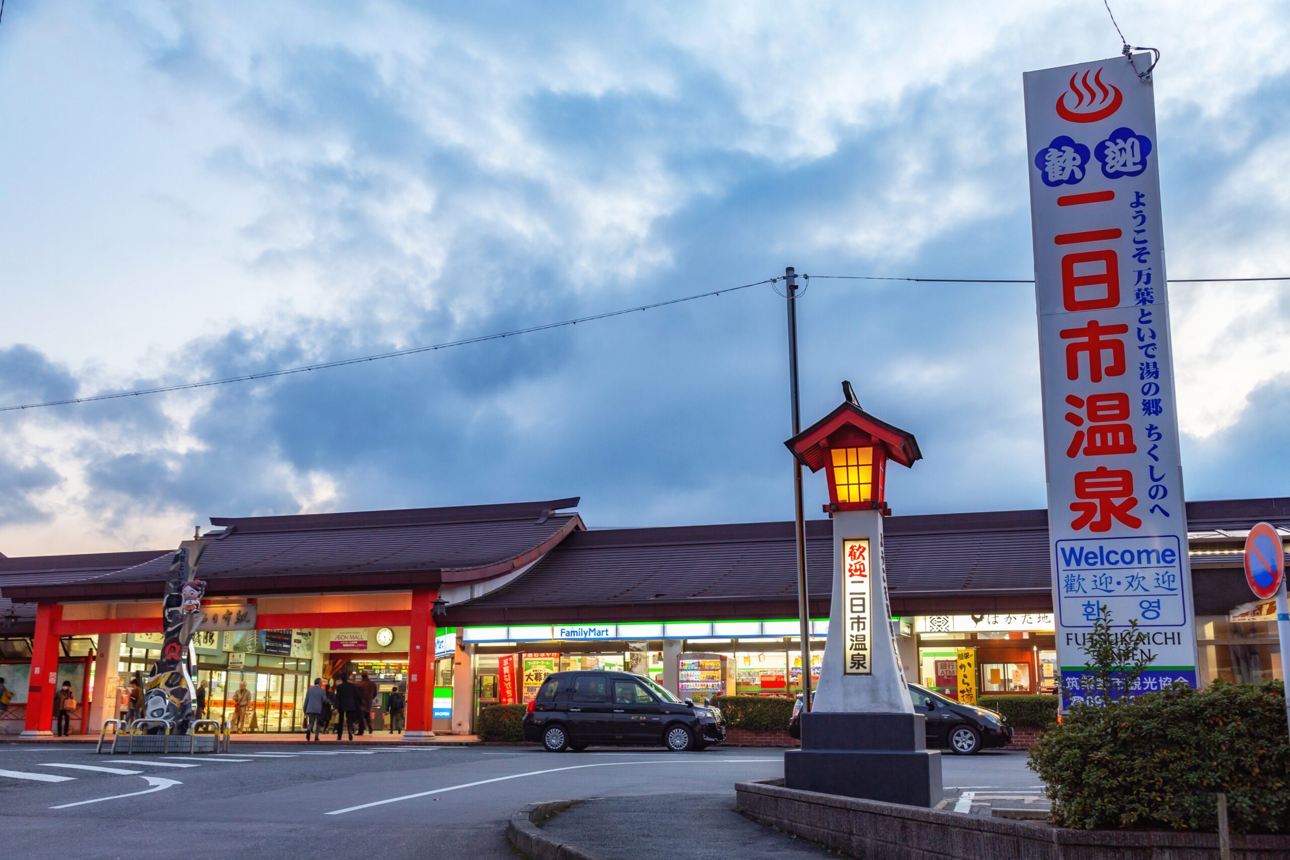 Front view of a Japanese shopping street with a tall white sign featuring red and blue Japanese characters, a red lantern, and storefronts including FamilyMart under a cloudy sky.