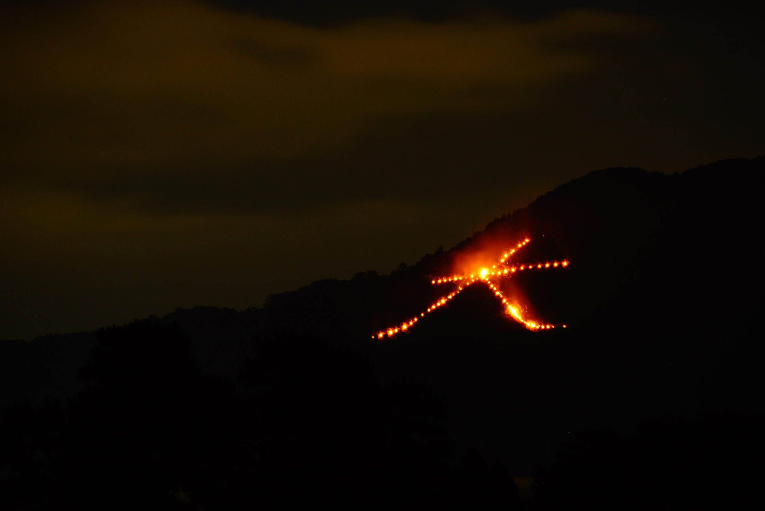 Night scene of a hillside with a zigzag line of orange lights or torches along a slope, glowing against a dark sky.