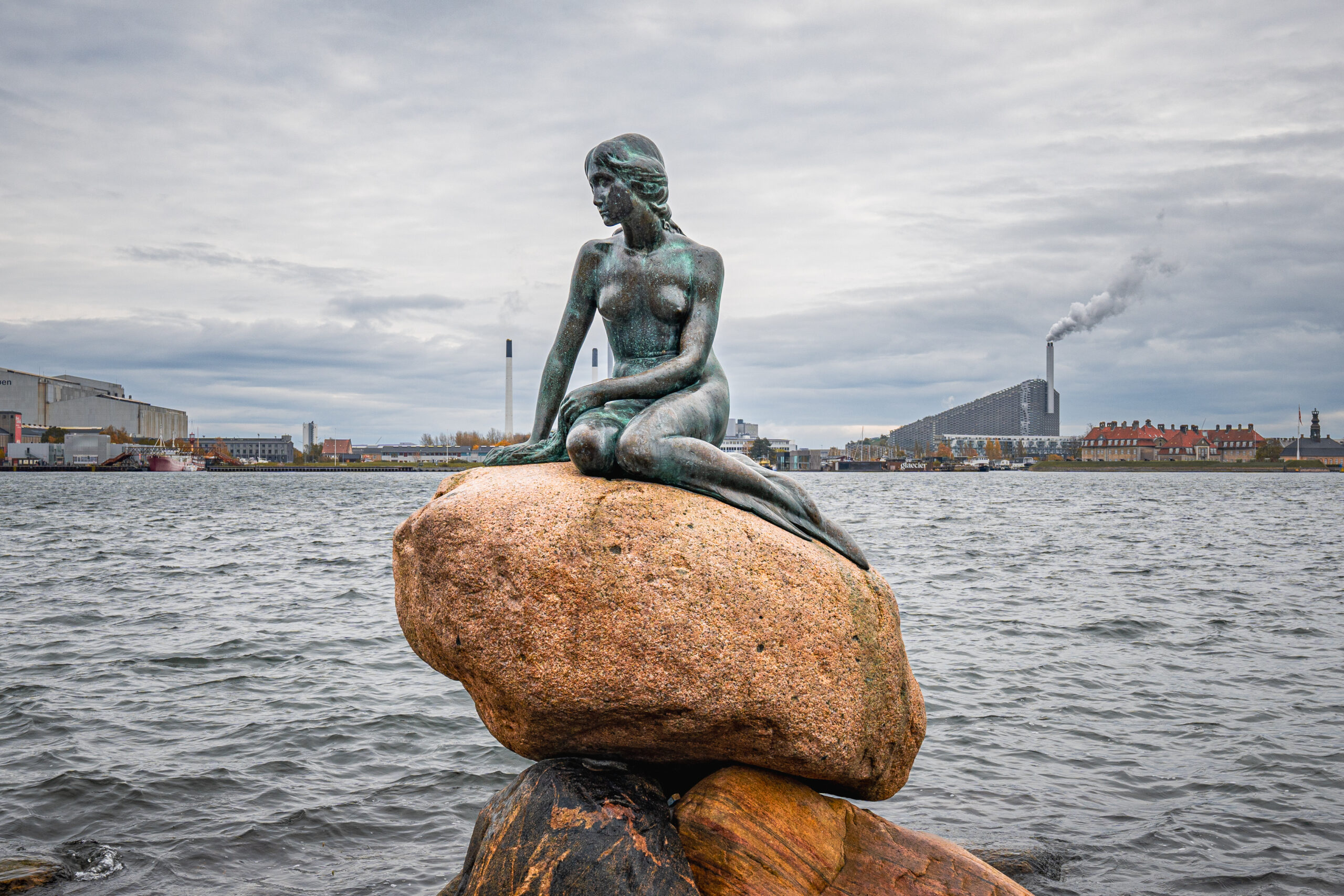 Bronze statue of a seated mermaid on a rocky outcrop by a harbor with industrial buildings and smokestacks in the background.