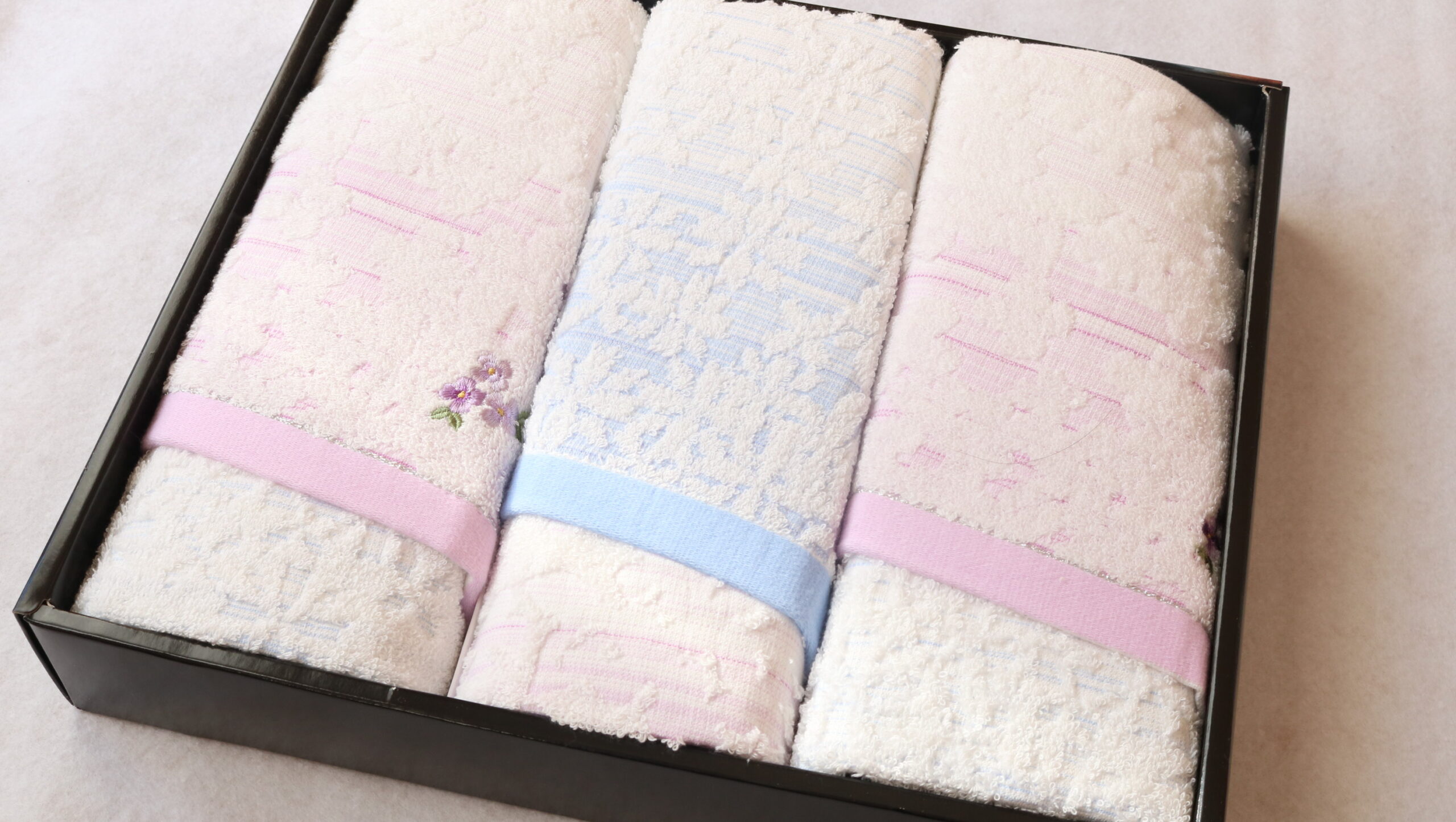 Set of three pastel towels—pink, blue, and white lace—rolled and placed in a black gift box with ribbons.