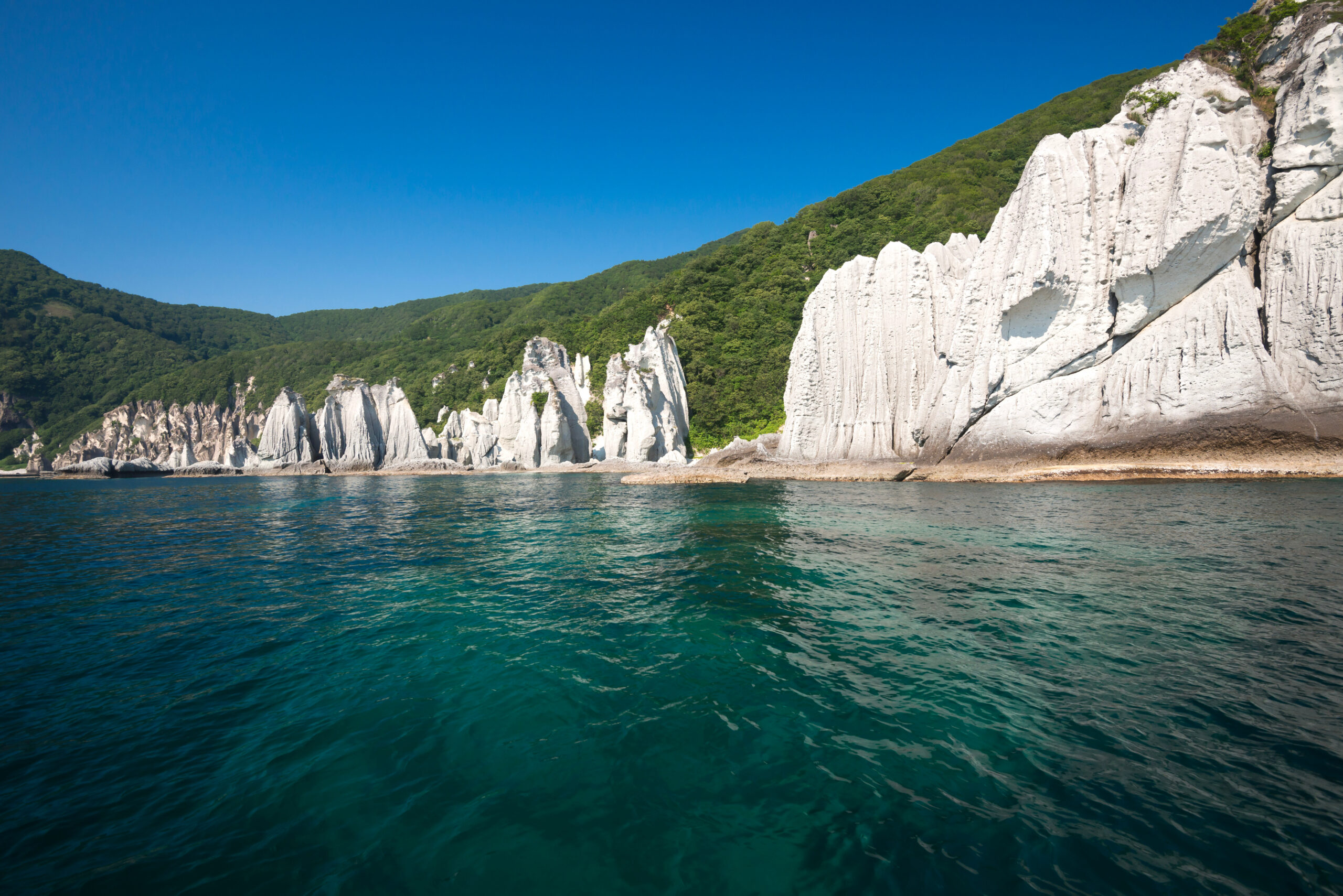 White chalk cliffs along a forested coastline under a bright blue sky, with calm teal water in the foreground.