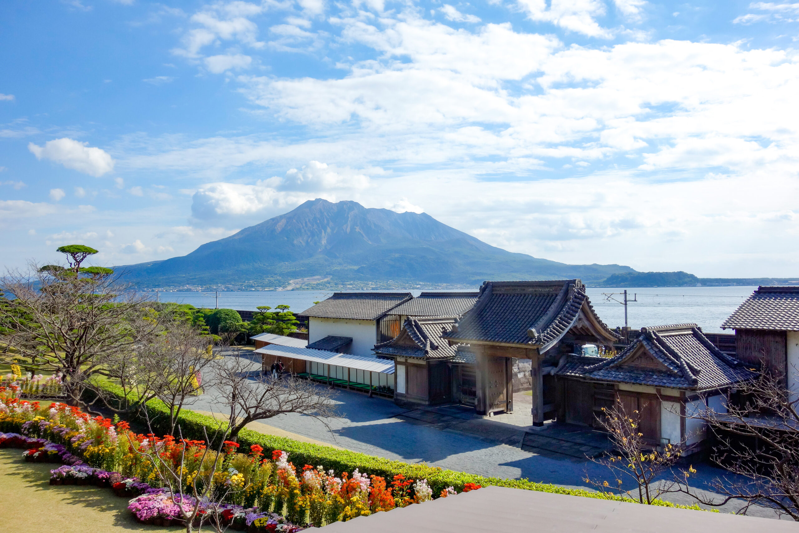 Colorful flower garden in the foreground with traditional Japanese buildings, a lake, and a distant mountain under a blue sky.