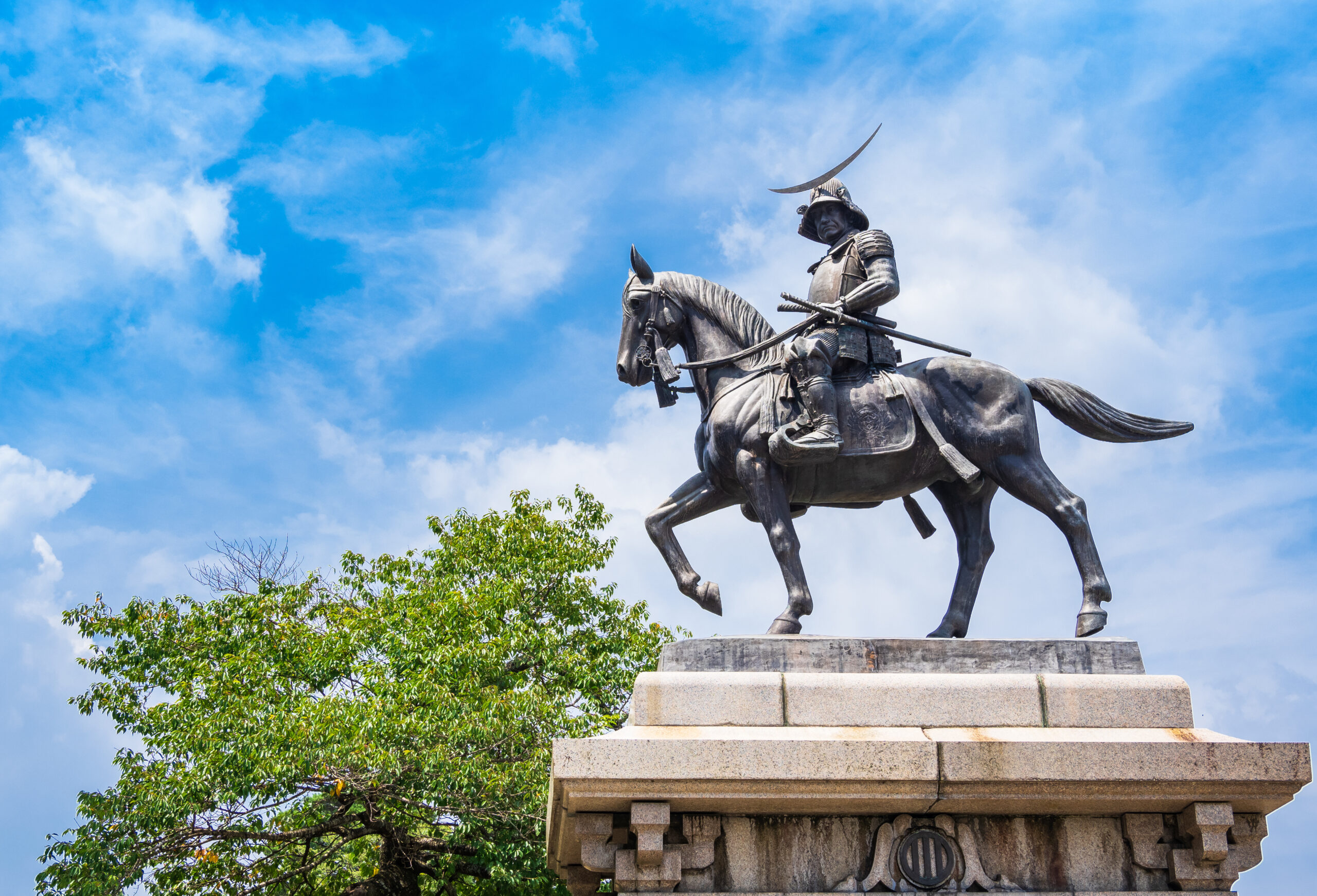 Bronze statue of a armored samurai on a horse atop a stone pedestal against a blue sky.