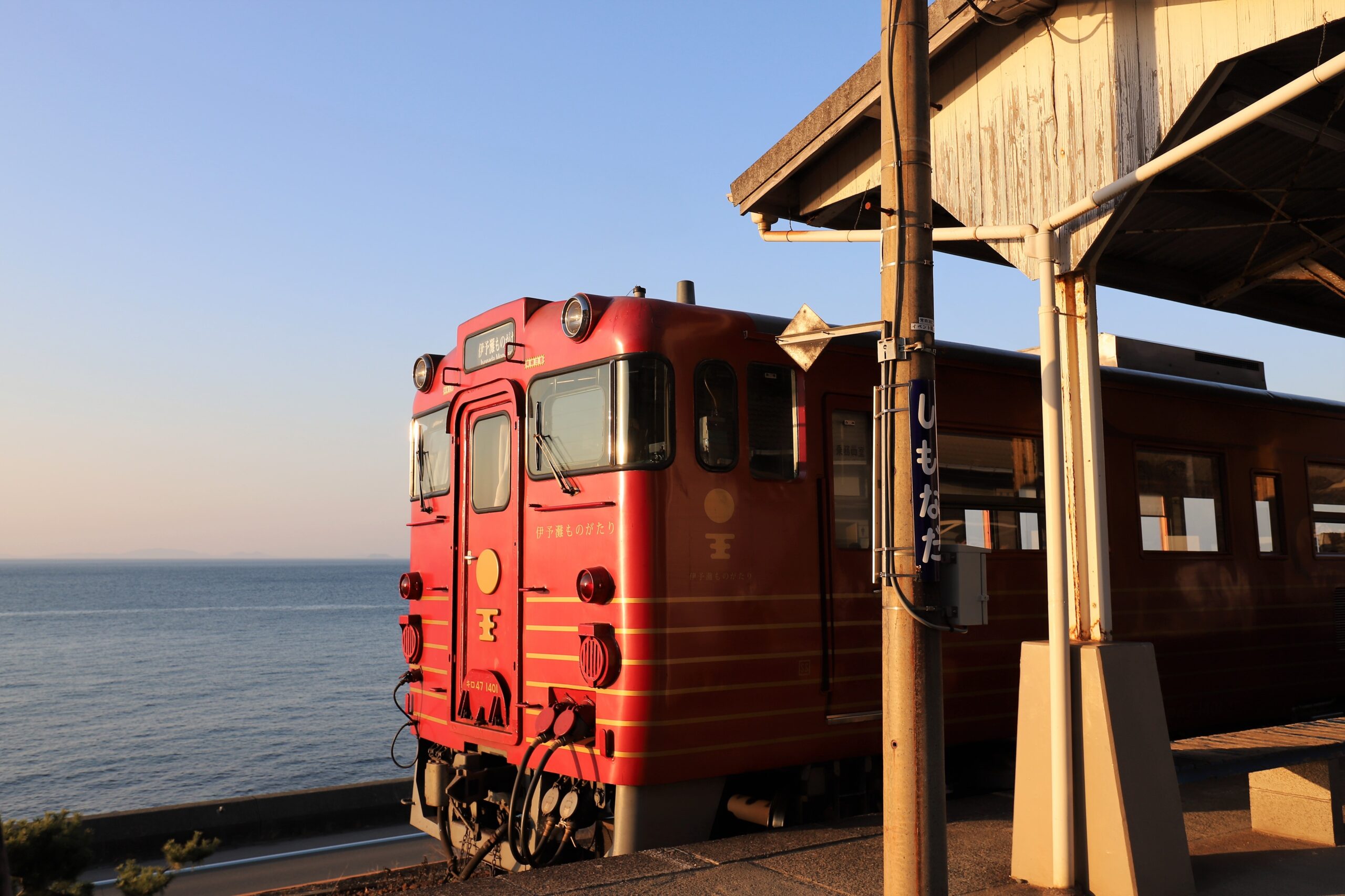Red locomotive parked at a coastal station, with rails and a weathered platform against a clear blue sky and sea in the background.